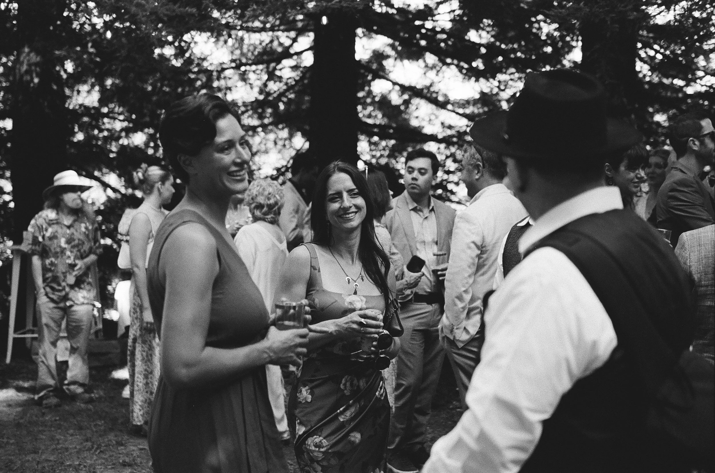 People gathered outdoors at a social event, talking and holding drinks, with trees in the background.