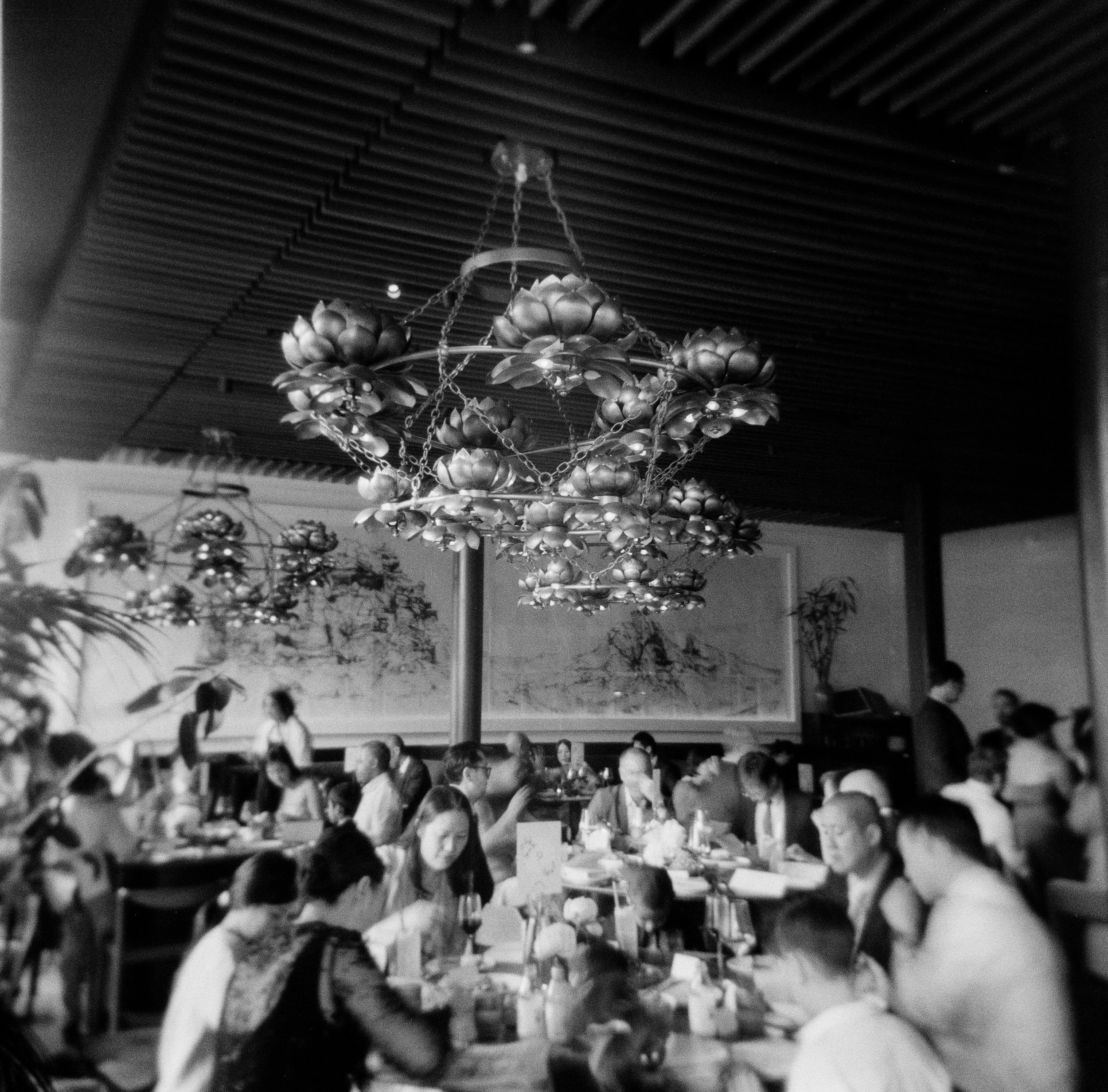 A black and white Holga photo of a dinner reception at Mister Jiu's with guests eating at a dining table with a chandelier hanging from the ceiling above them.