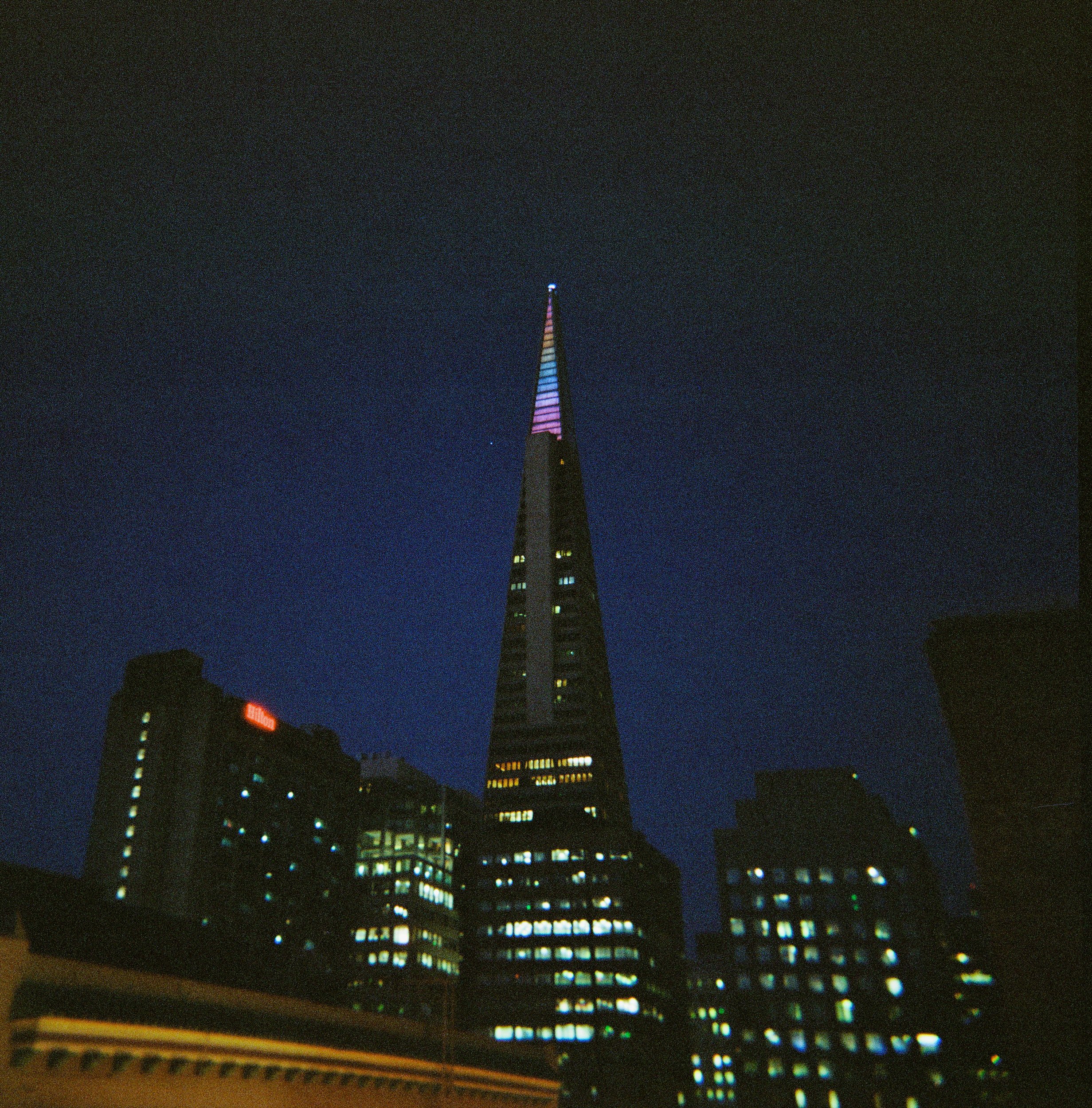 Nighttime cityscape featuring a tall, illuminated skyscraper with multicolored lights.