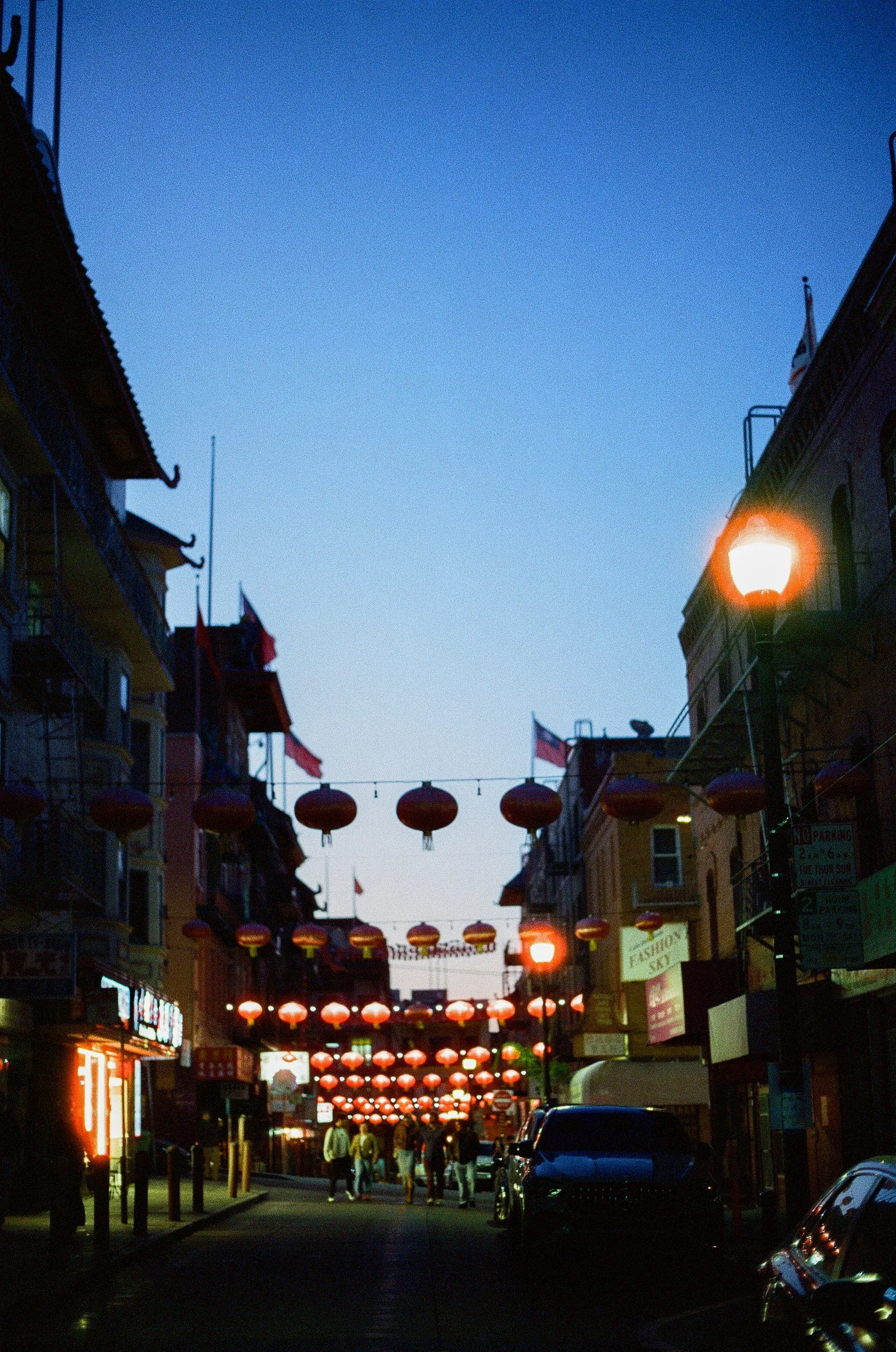 A busy street scene at dusk with buildings on both sides, red lanterns hanging across the street, and people walking on the sidewalk.