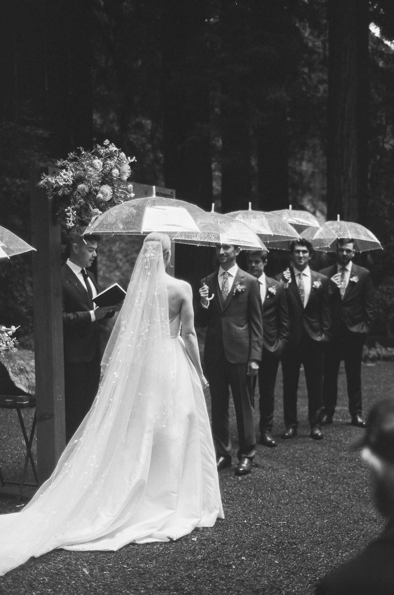 A wedding ceremony outdoors with a bride in a wedding dress and veil, holding a bouquet of flowers, standing under an umbrella with groomsmen in suits standing nearby.
