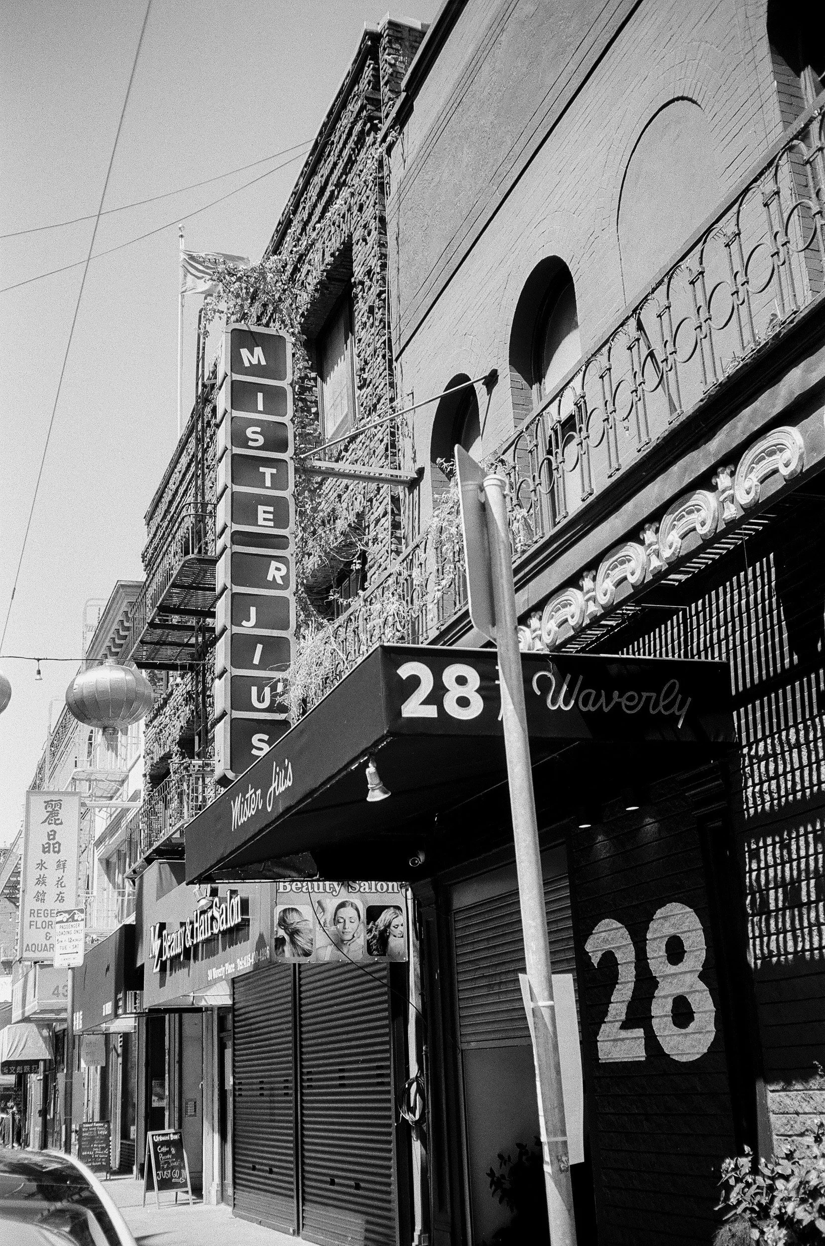 Street view of a building with a vertical sign reading 'Mister Jiu's.' and various storefront signs, including beauty and hair salons, with a sidewalk and hanging lanterns.