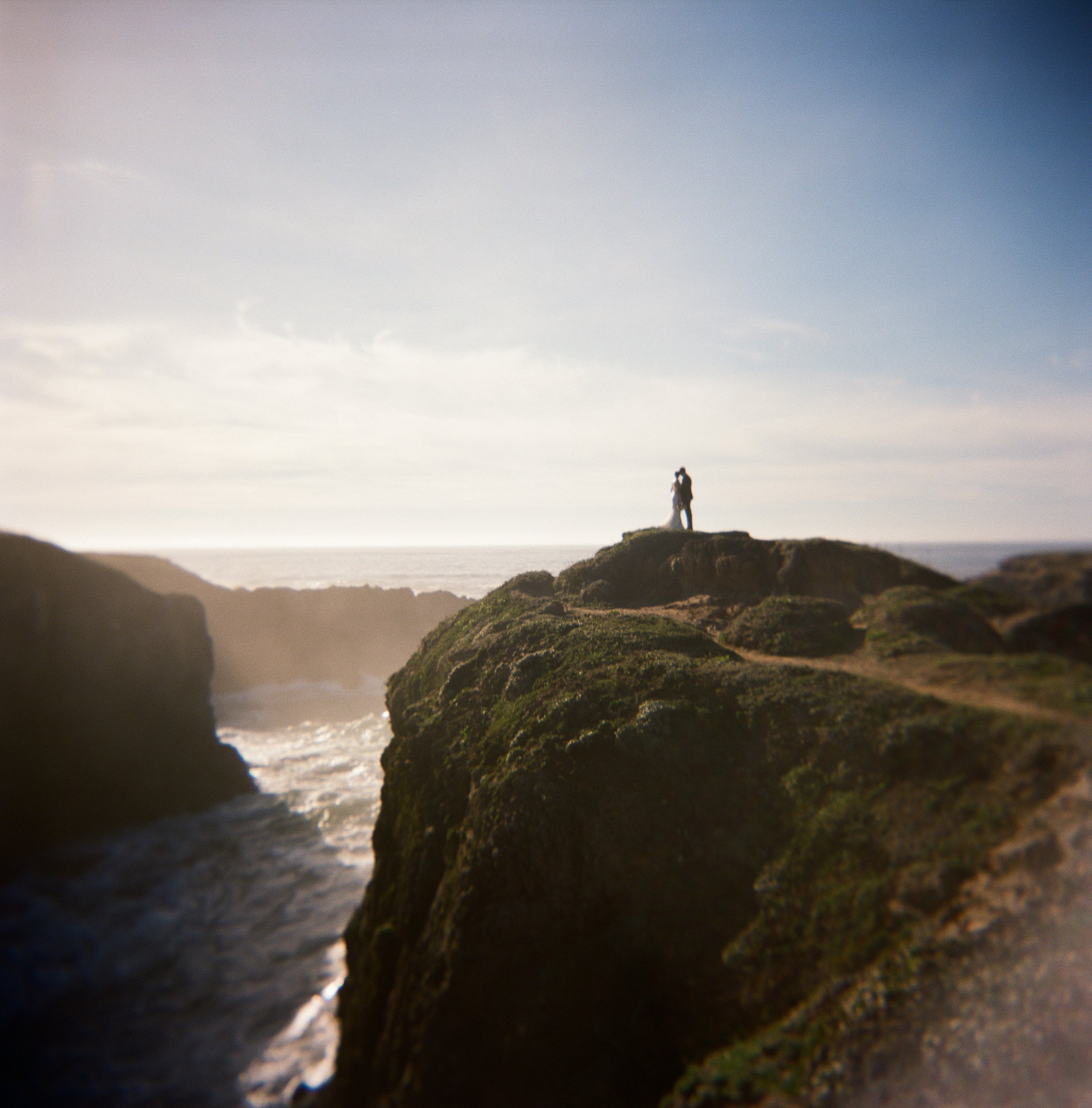 Two people standing on a small hilltop overlooking the ocean with rocky cliffs, under a clear blue sky.
