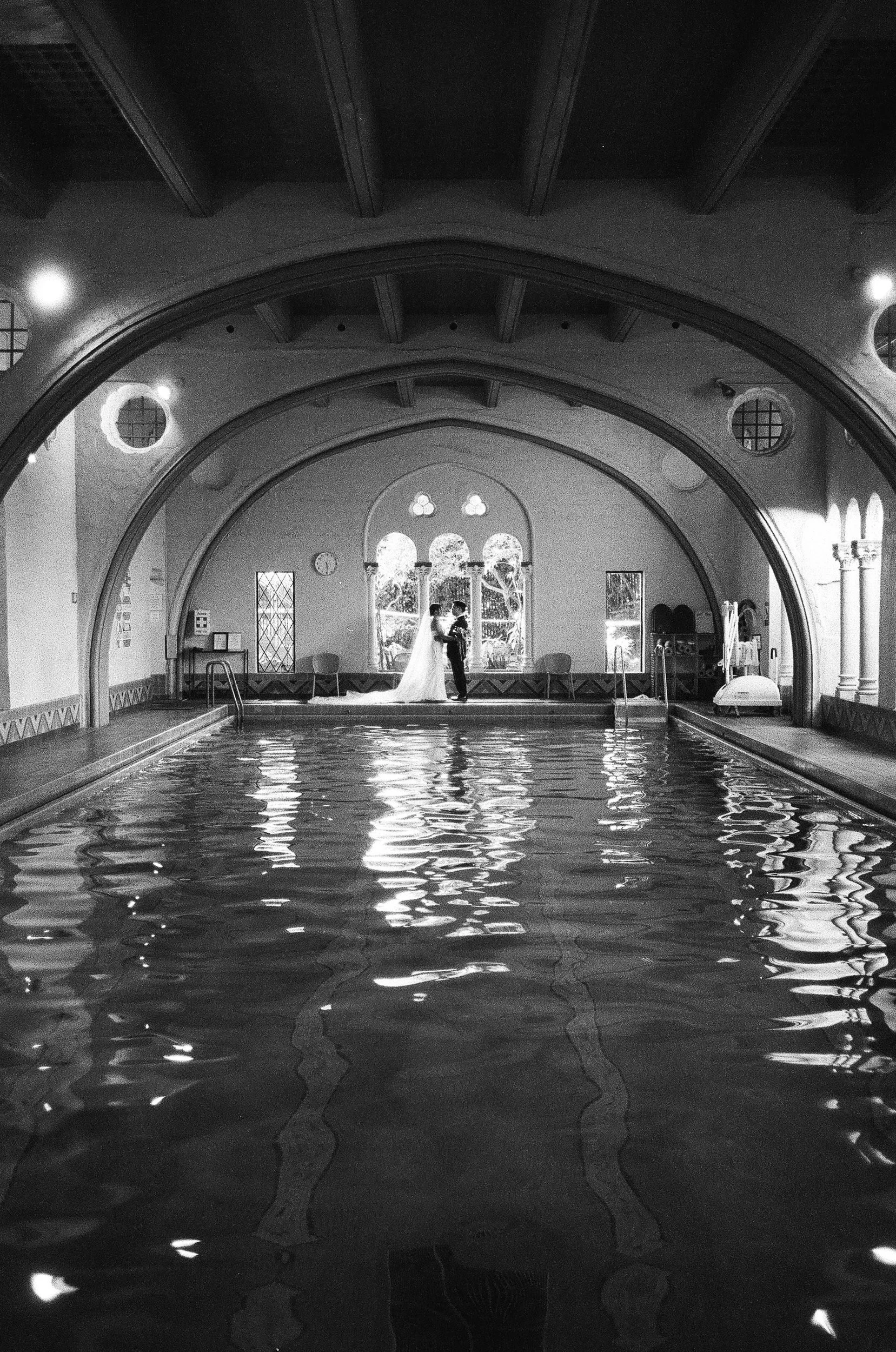 A bride and groom standing together in the center of an indoor swimming pool with arched windows behind them. They are holding hands, and the bride is wearing a wedding dress with a long train. The scene is reflected in the water, and the setting has