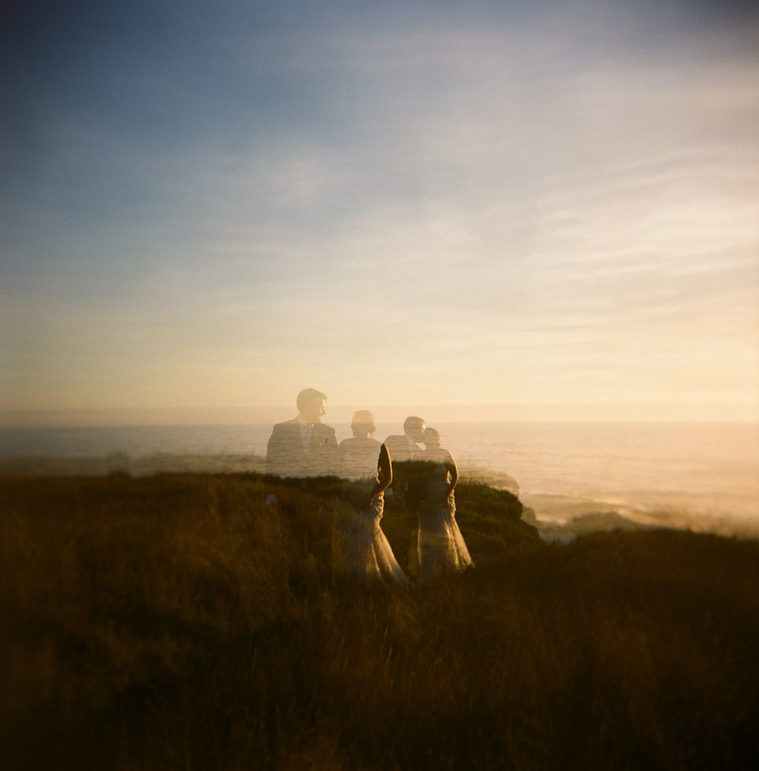A group of people standing on a grassy hill during sunset, overlooking the ocean with a clear sky above.