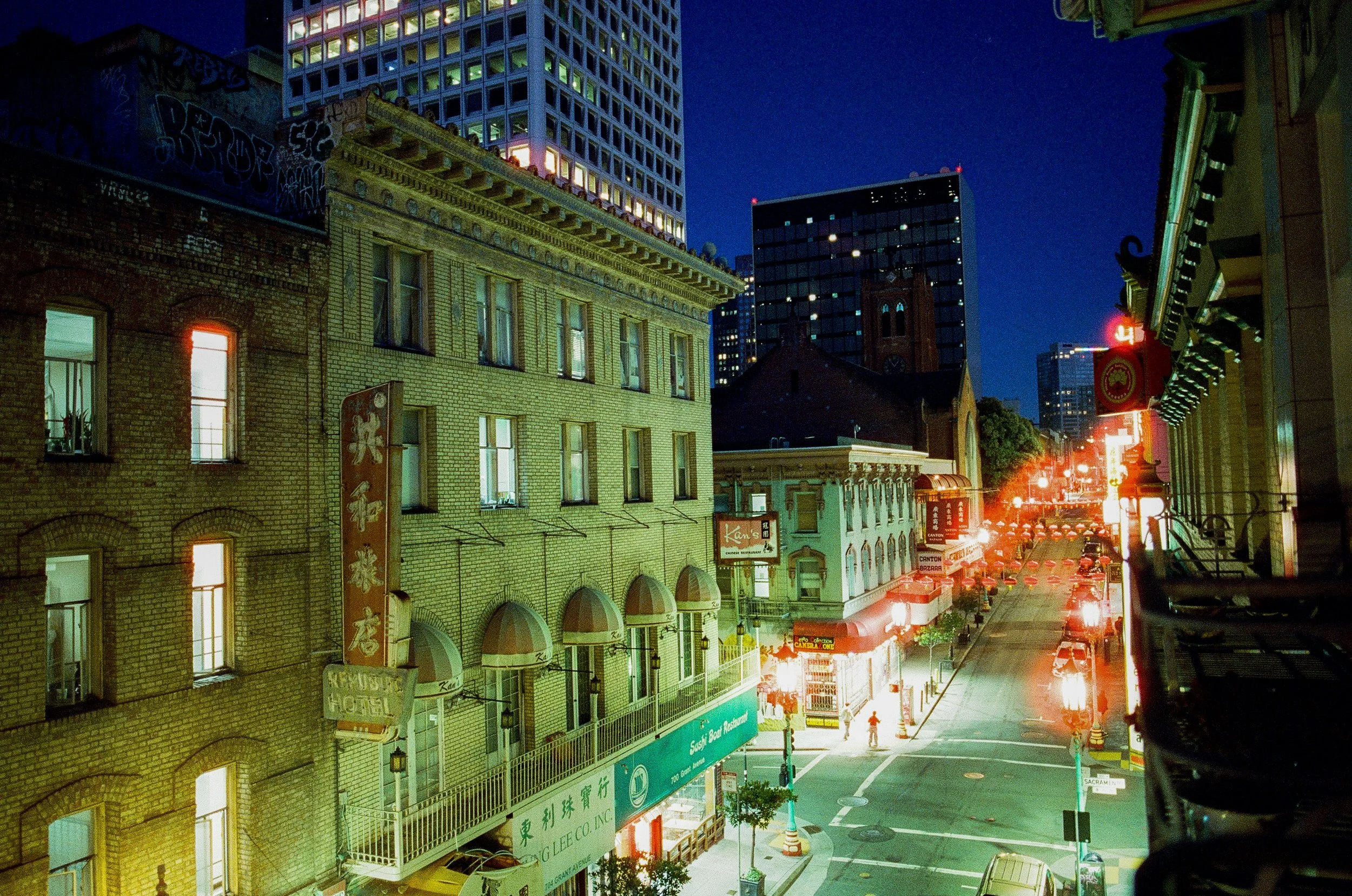Nighttime city street scene with multi-story buildings, neon signs, and cars parked along the street. A modern high-rise with lit windows contrasts with older brick buildings. There are street signs, storefronts, and a few pedestrians visible.