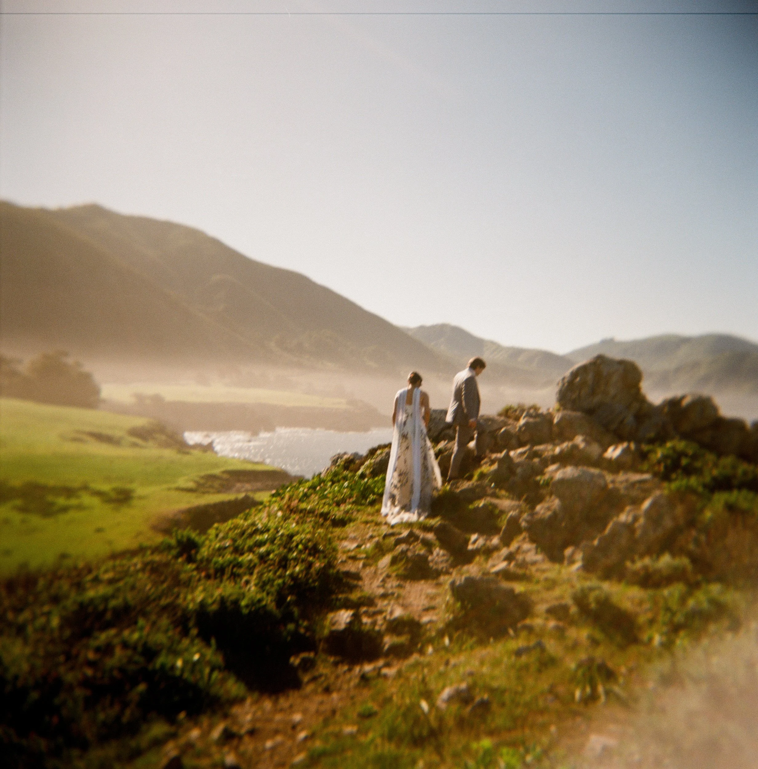A landscape scene with hills or dunes and a clear sky, and three people walking along a rocky path.