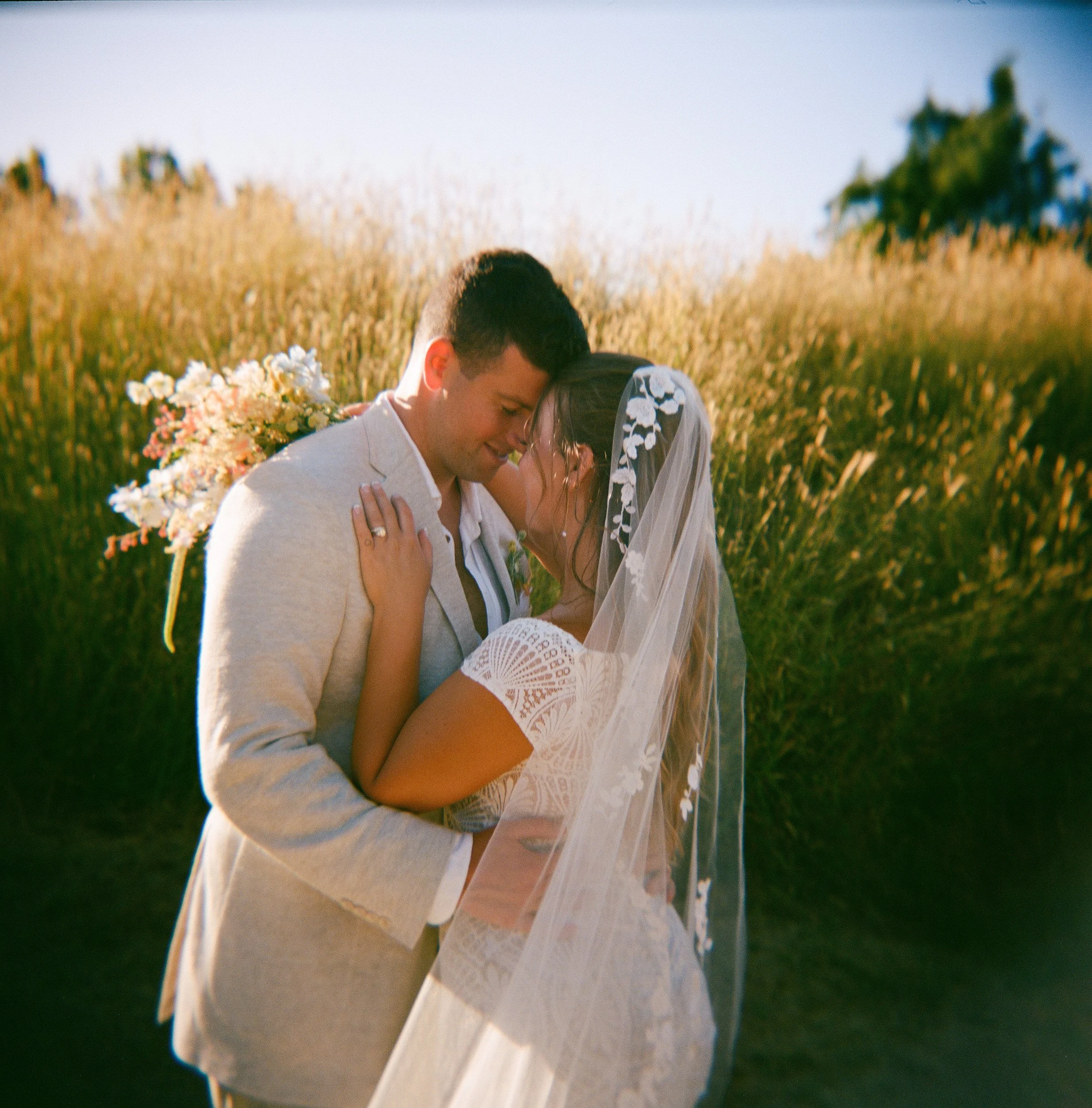 A couple kissing outdoors in a field during daylight, with a bouquet of flowers nearby.