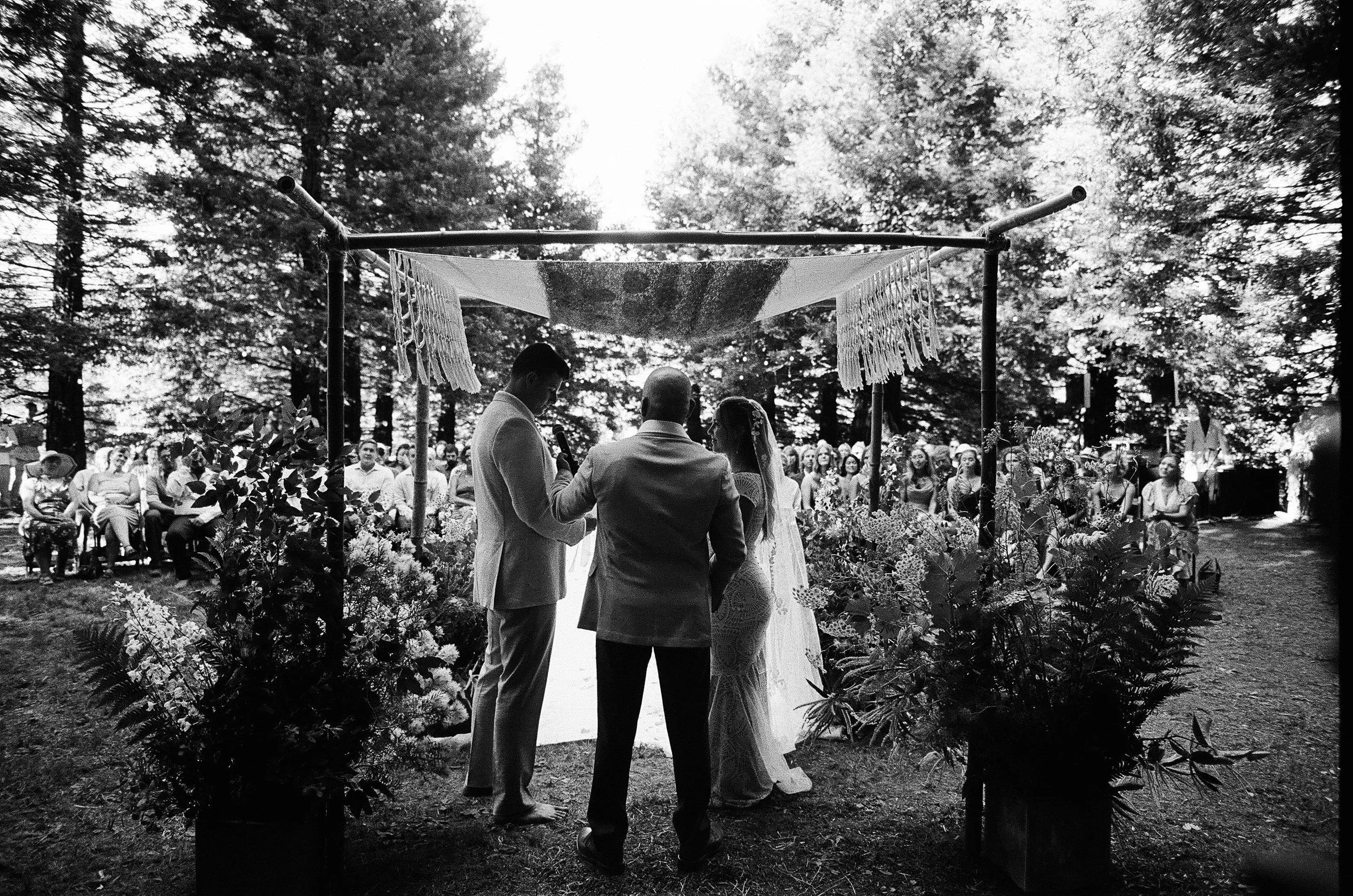 Black and white photo of a wedding ceremony outdoors in a wooded area. The bride and groom stand facing each other under a canopy with floral arrangements, while the officiant conducts the ceremony. Guests sit and watch in the background.