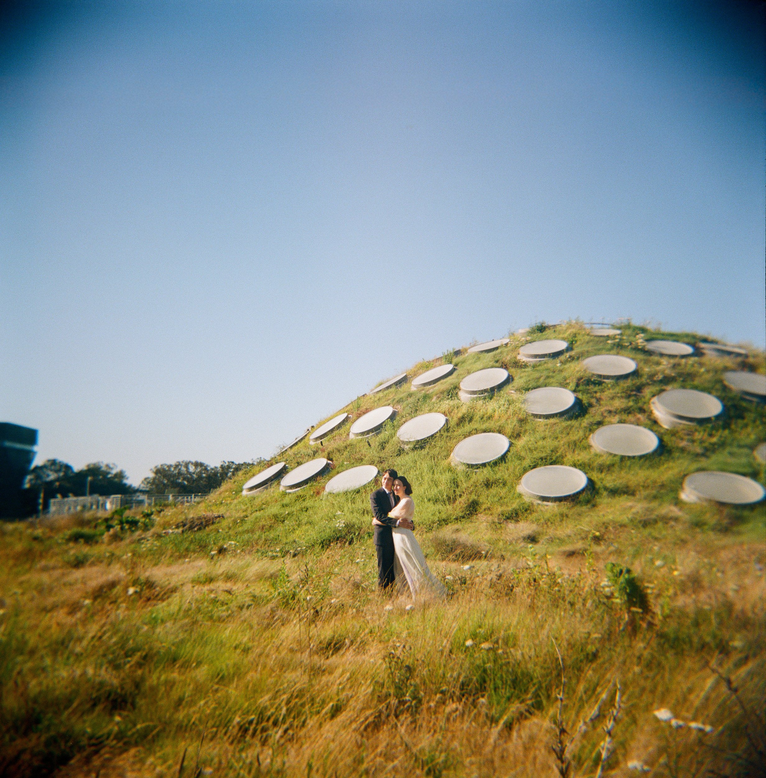 A couple in wedding attire standing outdoors on a grassy hill, with a large hill covered in numerous satellite dishes in the background.