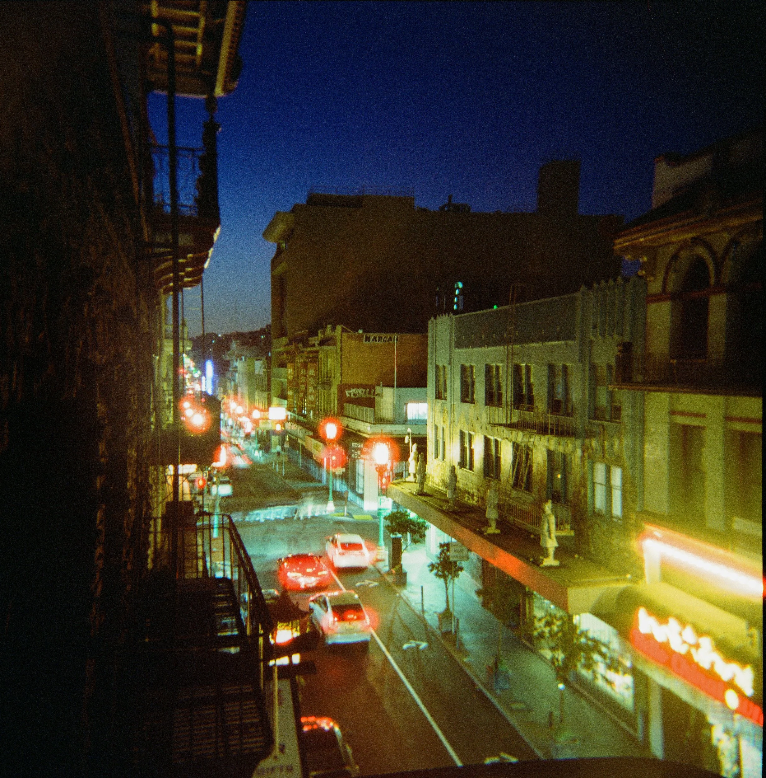 Nighttime scene of a city street with vehicles and buildings, illuminated by streetlights and neon signs, viewed from a balcony or elevated position.