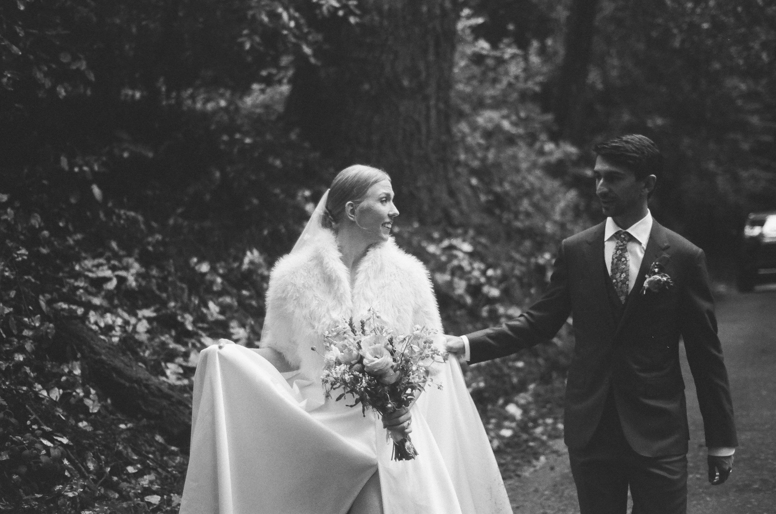 A bride and groom holding hands outdoors in a wooded area, with the bride wearing a white dress and a fur stole, and the groom in a dark suit and floral tie.