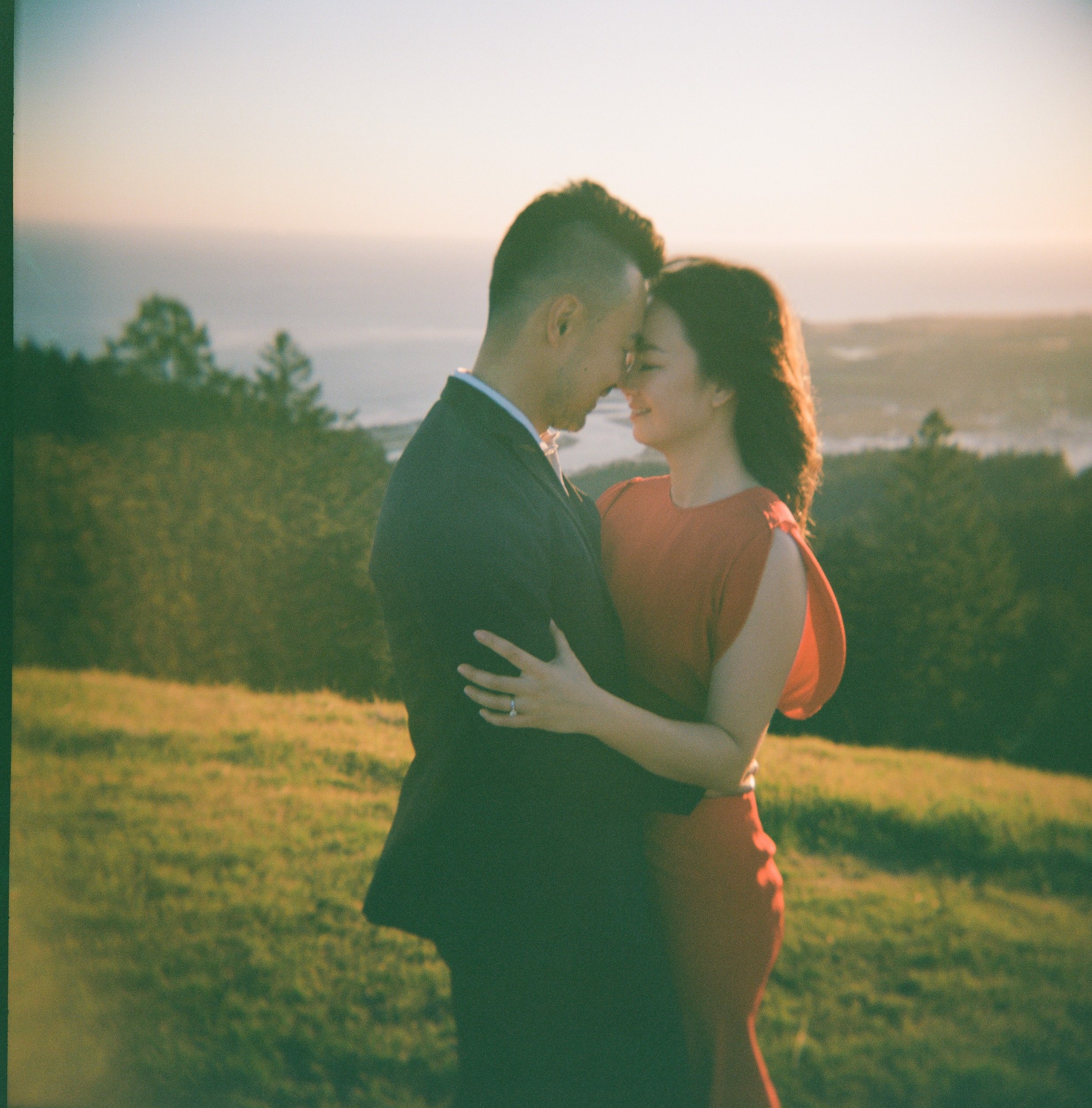 A couple is standing close together outdoors during sunset or sunrise, with the ocean and a few trees in the background. They are facing each other with closed eyes, about to kiss or sharing an intimate moment.