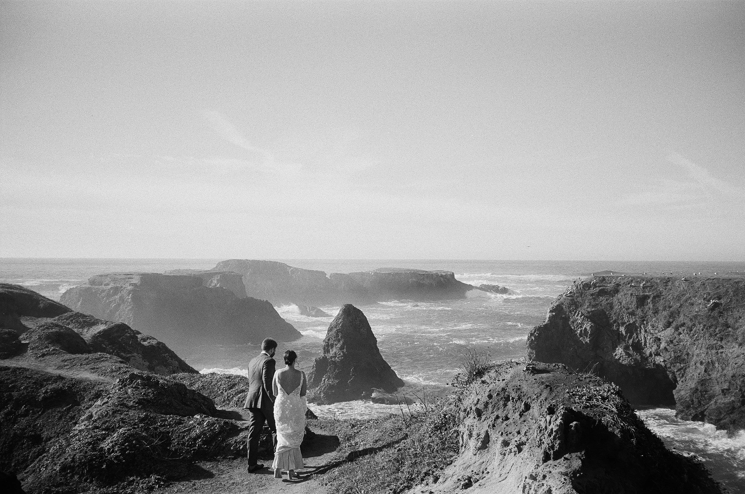 A black and white photo of a couple holding hands on a rocky beach, looking out at the ocean with large rocks and cliffs in the background.