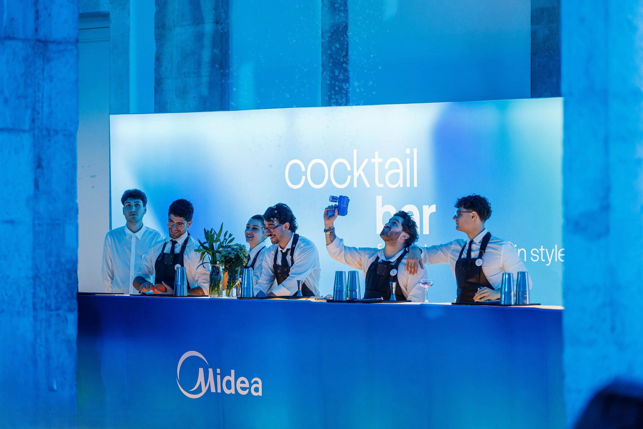 Equipe de bartenders sorridentes na bancada do bar durante evento de coquetel, com um fundo com as palavras "cocktail bar" e a marca Midea, sob iluminação azul.