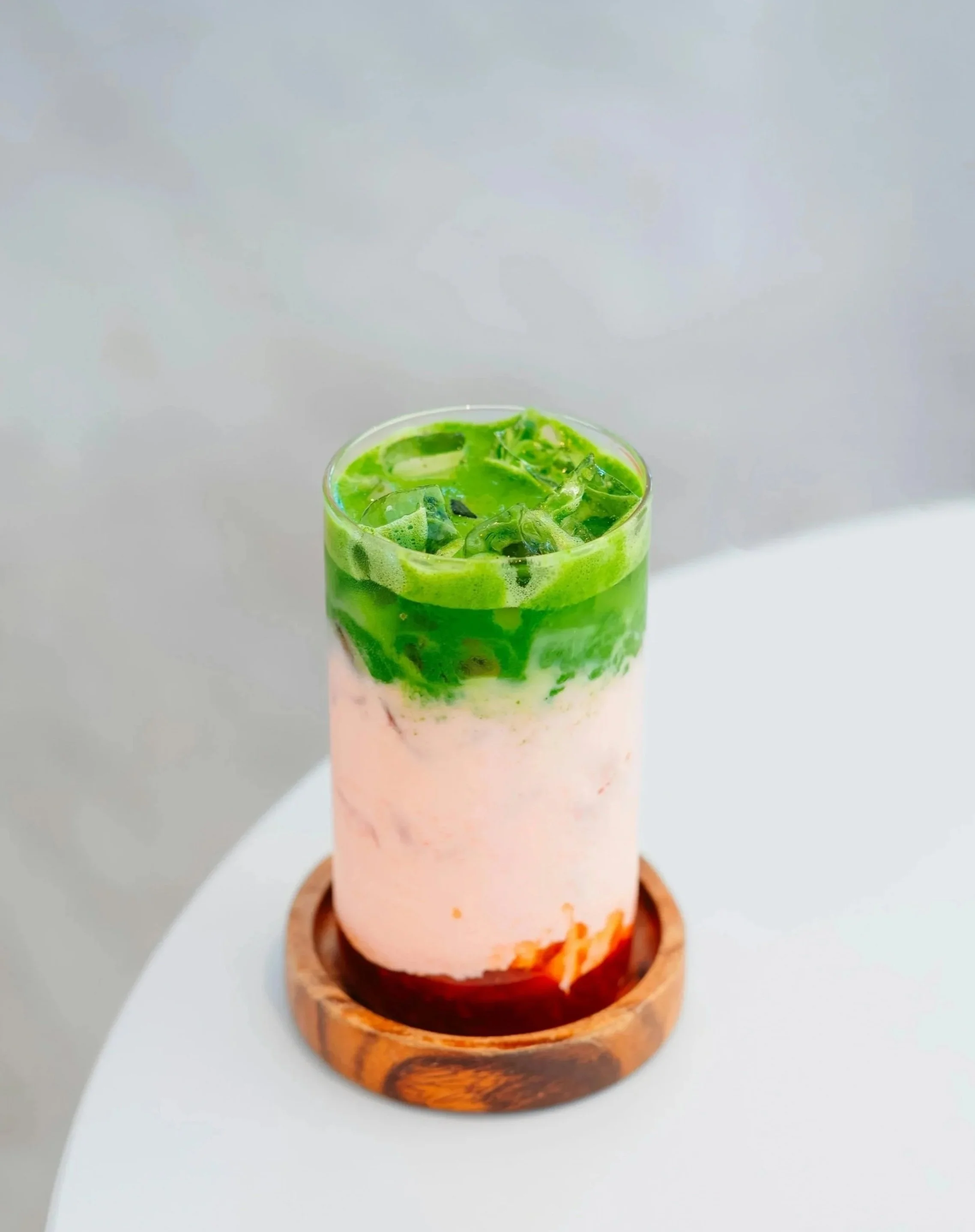 Energy Bar, Copo de smoothie com camadas de frutas, incluindo morango na base, creme de leite no meio e cobertura de chá verde com gelo no topo, servido em um suporte de madeira.