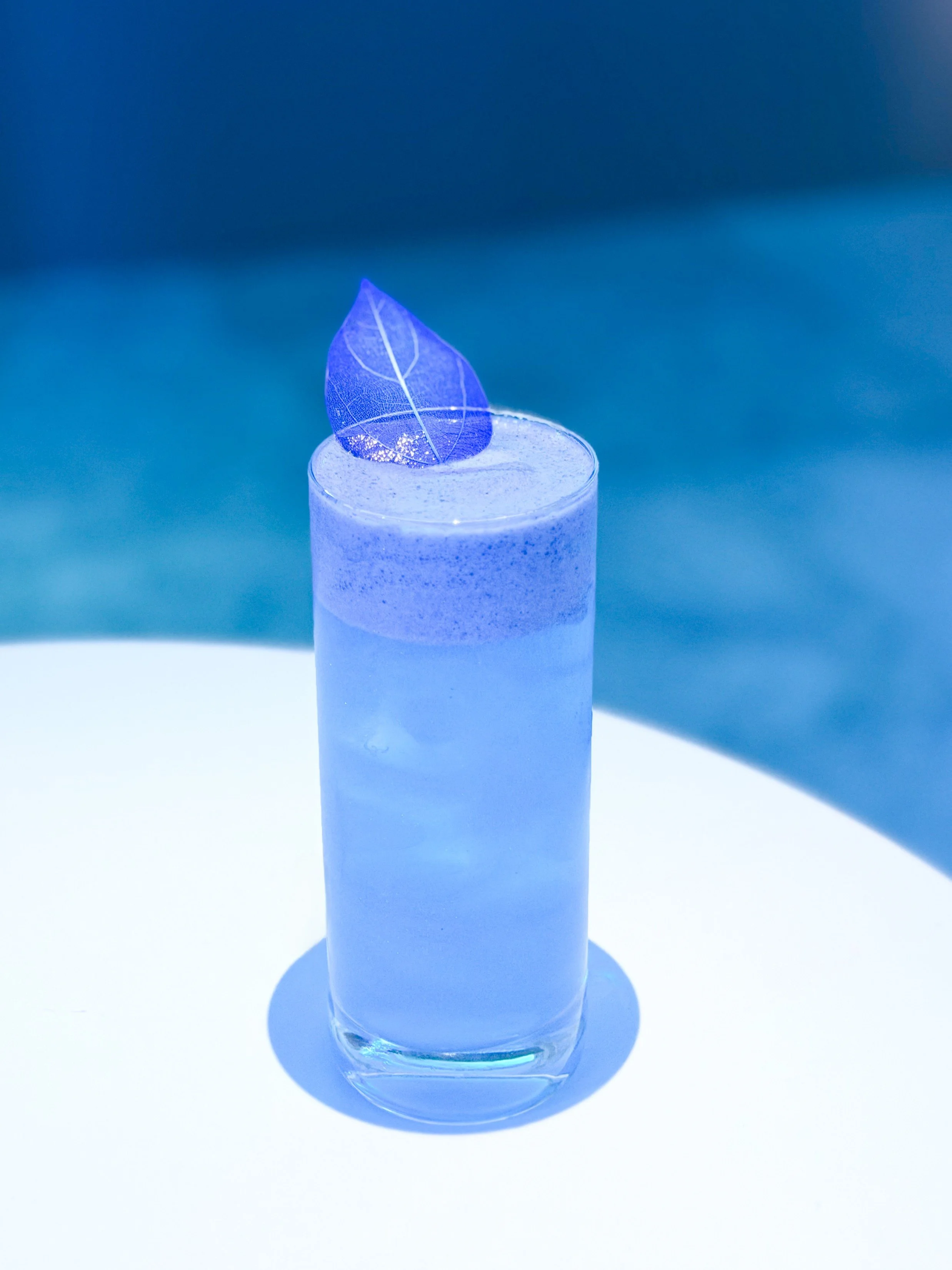 Cocktail branding em copo alto com bebida azul, decorado com uma folha azul transparente por cima.