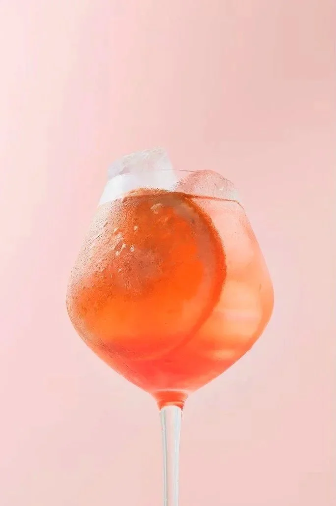 Copo de drink com gelo, uma fatia de laranja e um pavio, contra fundo rosa claro.