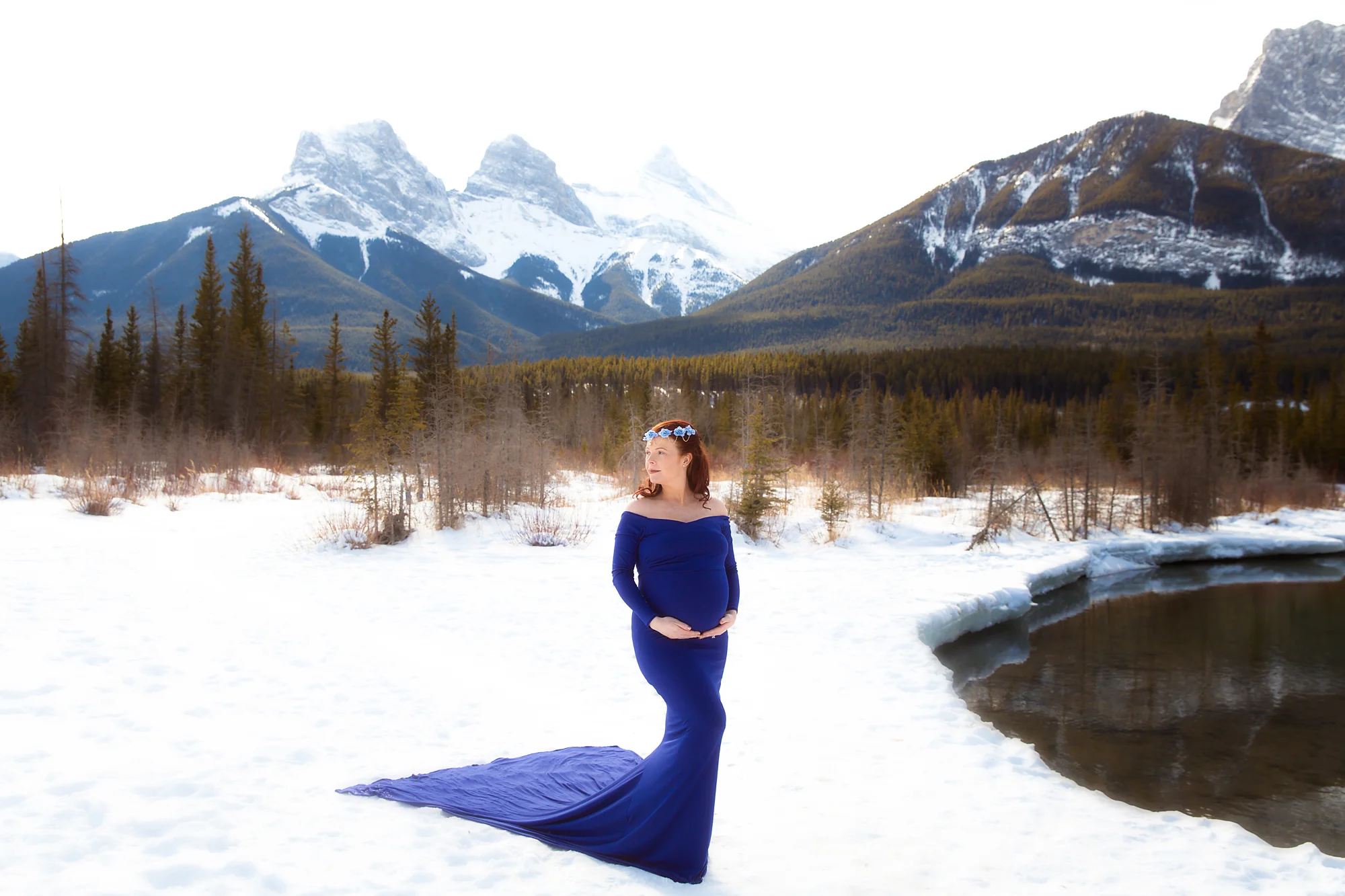 Beautiful mother-to-be in a gorgeous royal blue gown standing in front of The Three Sister mountains in Canmore, Alberta, Canada during winter. Calgary, Airdrie, Canmore, Banff and Kananaskis Maternity Photographer - Milashka photography