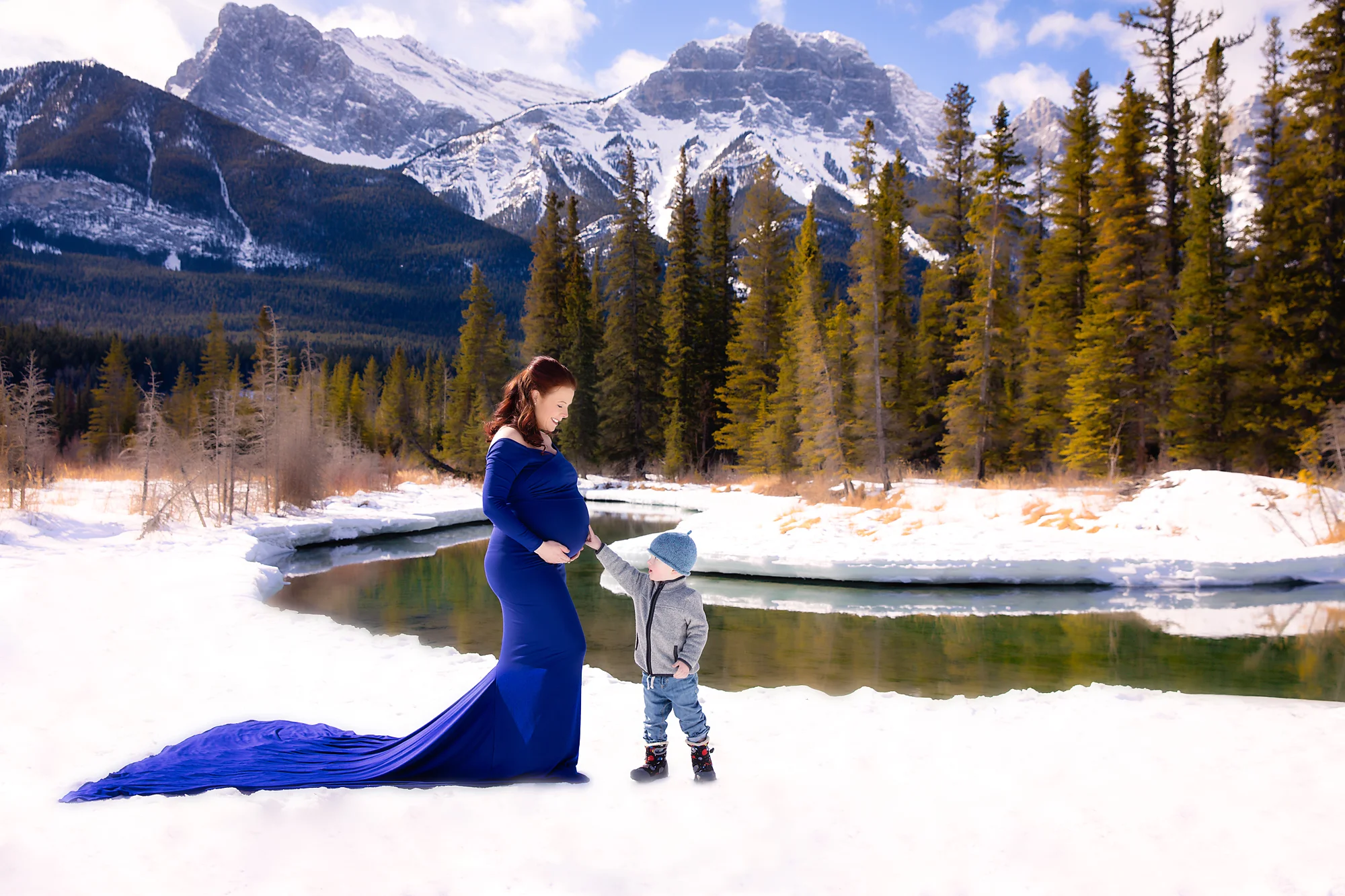 A little boy is touching his expecting mother’s belly. Mountain maternity session. Mom-to-be is wearing a beautiful royal blue gown and is standing on the snow in front of mountains and forest. Calgary Maternity photographer - Milashka Photography