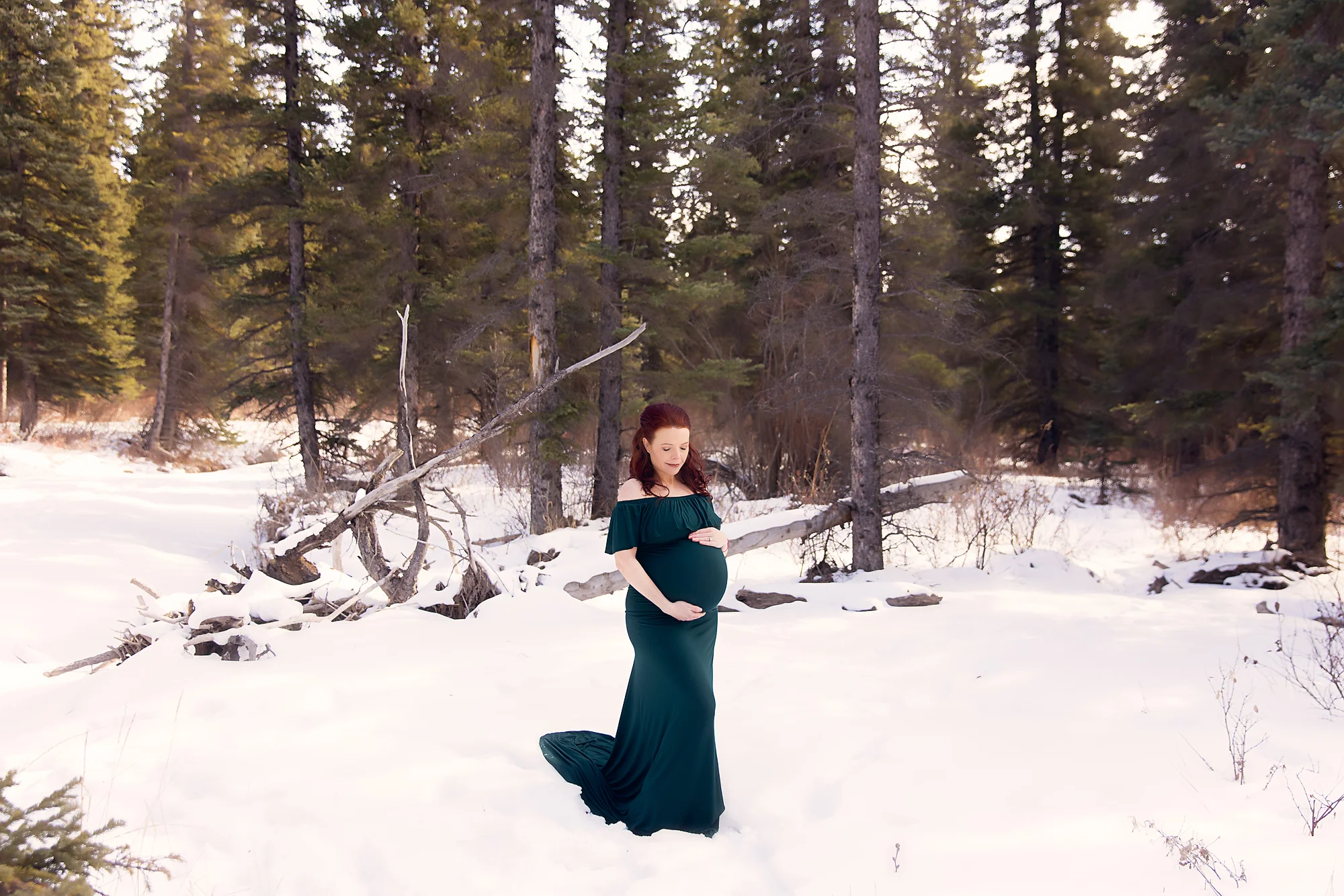 Expecting mother is wearing a green gown in the forest, standing on a snow. Maternity photoshoot ideas. Calgary Photographer - Milashka Photography