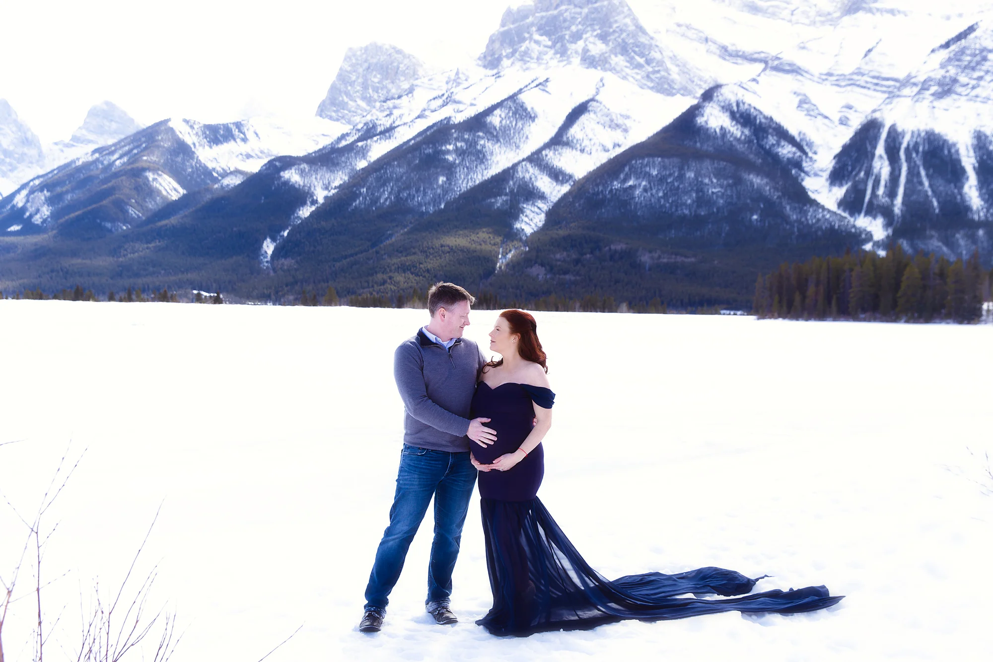 Mom and Dad to be standing on a snow in front of Rocky Mountains. Mom-to-be is wearing a gorgeous blue gown. Mountain Maternity Session in Kananaskis Country. Milashka Photography