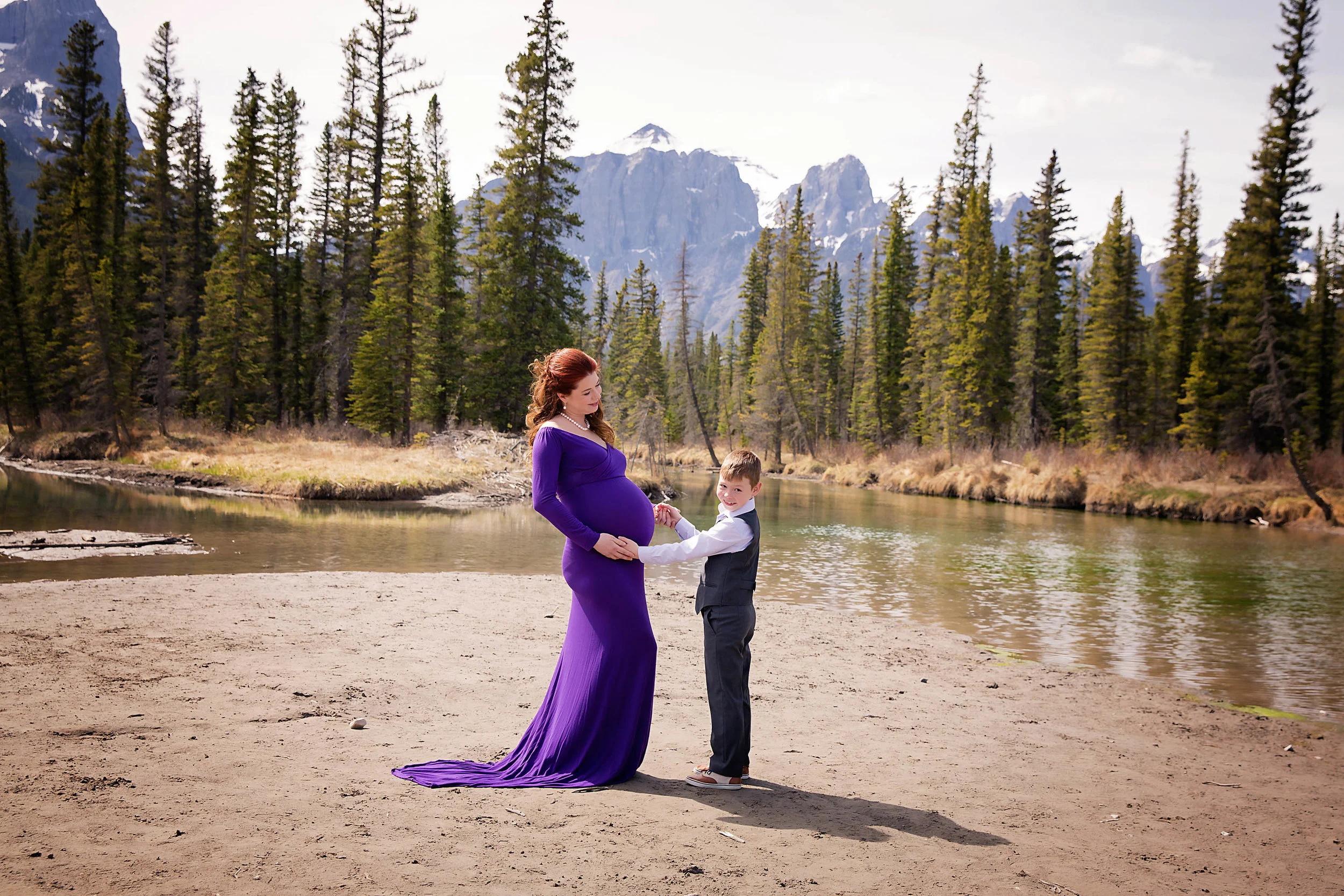 Mother and son maternity in the mountains shot. Mom to be is wearing a purple gown and looking at her son while a 7 year old boy is looking straight at the camera and smiling. Calgary maternity photographer - Milashka Photography