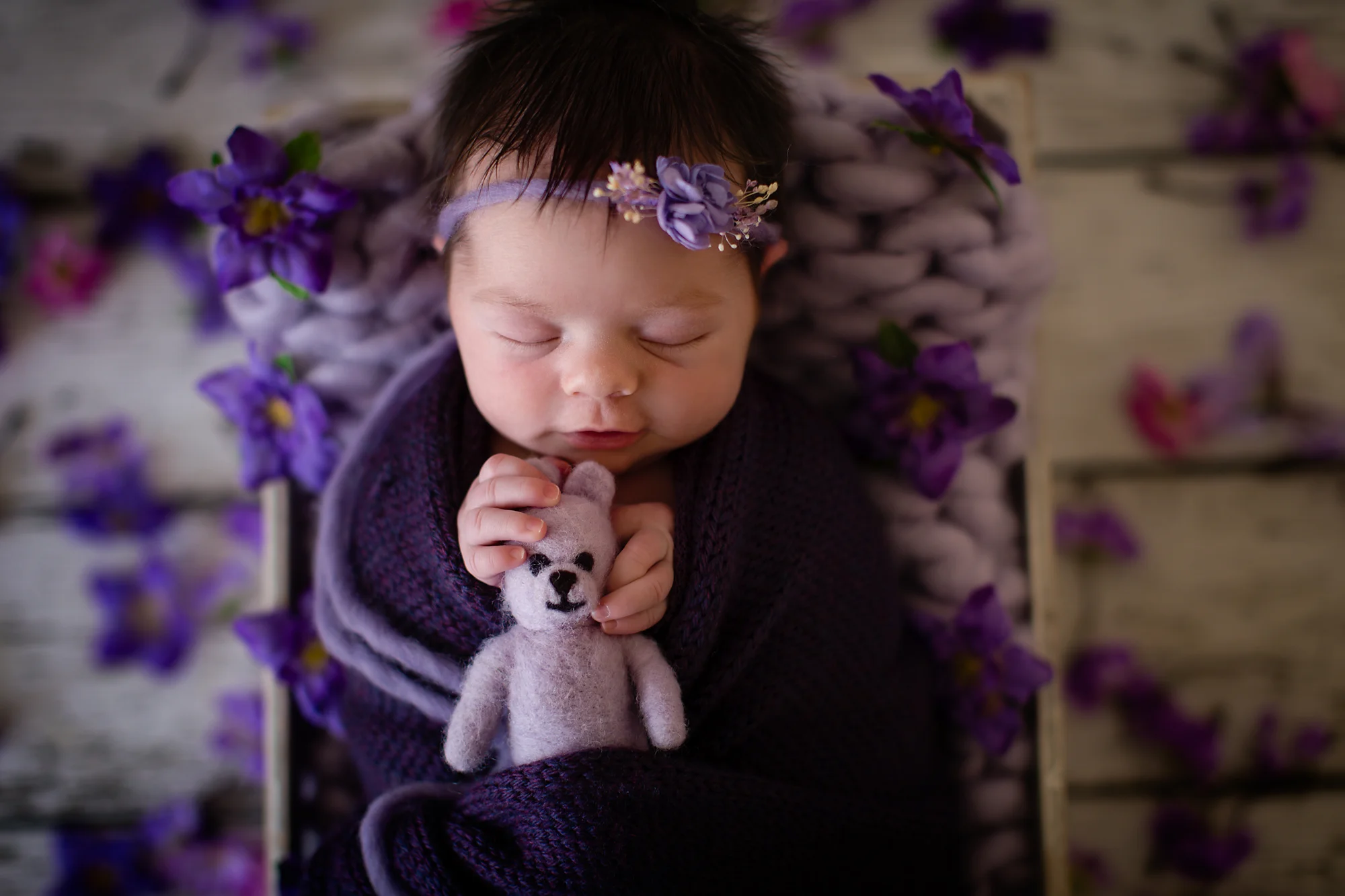 Newborn baby girl photoshoot idea. Baby girl is smiling in her sleep and is holding a little purple teddy bear. Posed in a crate with purple flowers around her.