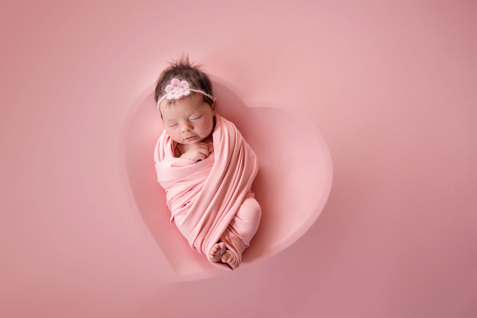 Newborn baby girl photoshoot idea. Baby girl is posed in a heart bowl covered by pink material. Calgary newborn photographer.