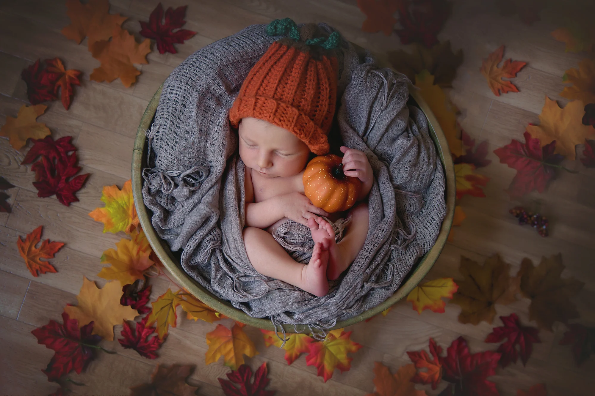 Thanksgiving inspired newborn baby photoshoot. Baby is dressed in a pumpkin hat, holding a little pumpkin, surrounded by leafs and is sleeping soundly in a wooden bowl. Newborn Photoshoot ideas. Calgary and Airdrie newborn baby photographer - Milashka Photography