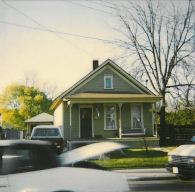 Small green single-story house with a front porch, a large leafless tree behind it, and blurred cars passing on the street in the foreground.