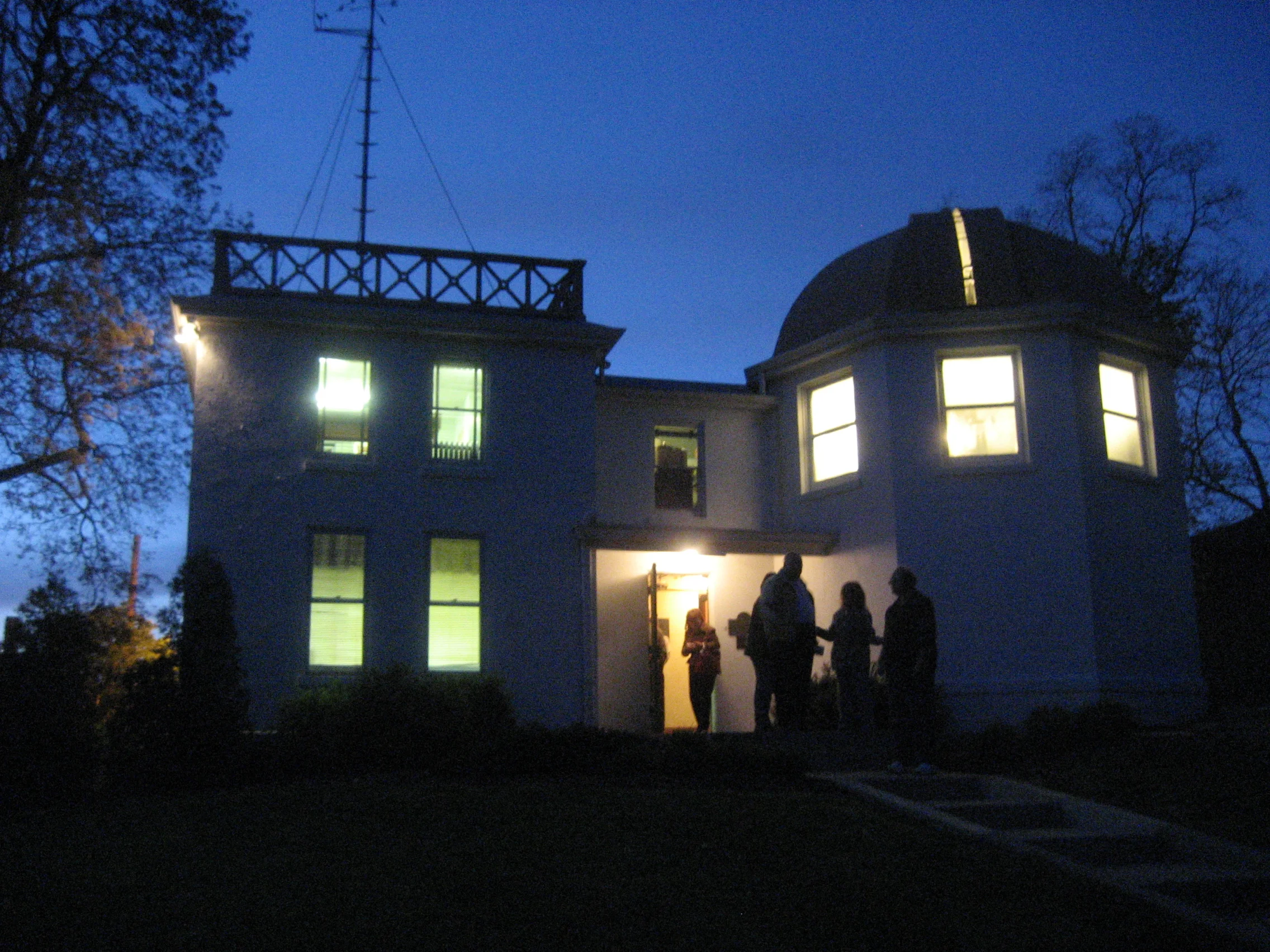 Two-story house at dusk with lights on and a group of people standing at the front door.