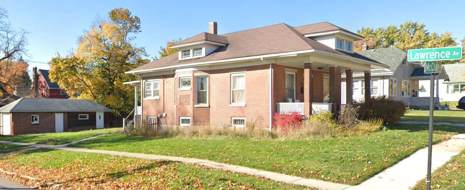 Brick bungalow with front porch on a grassy corner lot in autumn, sidewalk and a green "Lawrence Av" street sign.