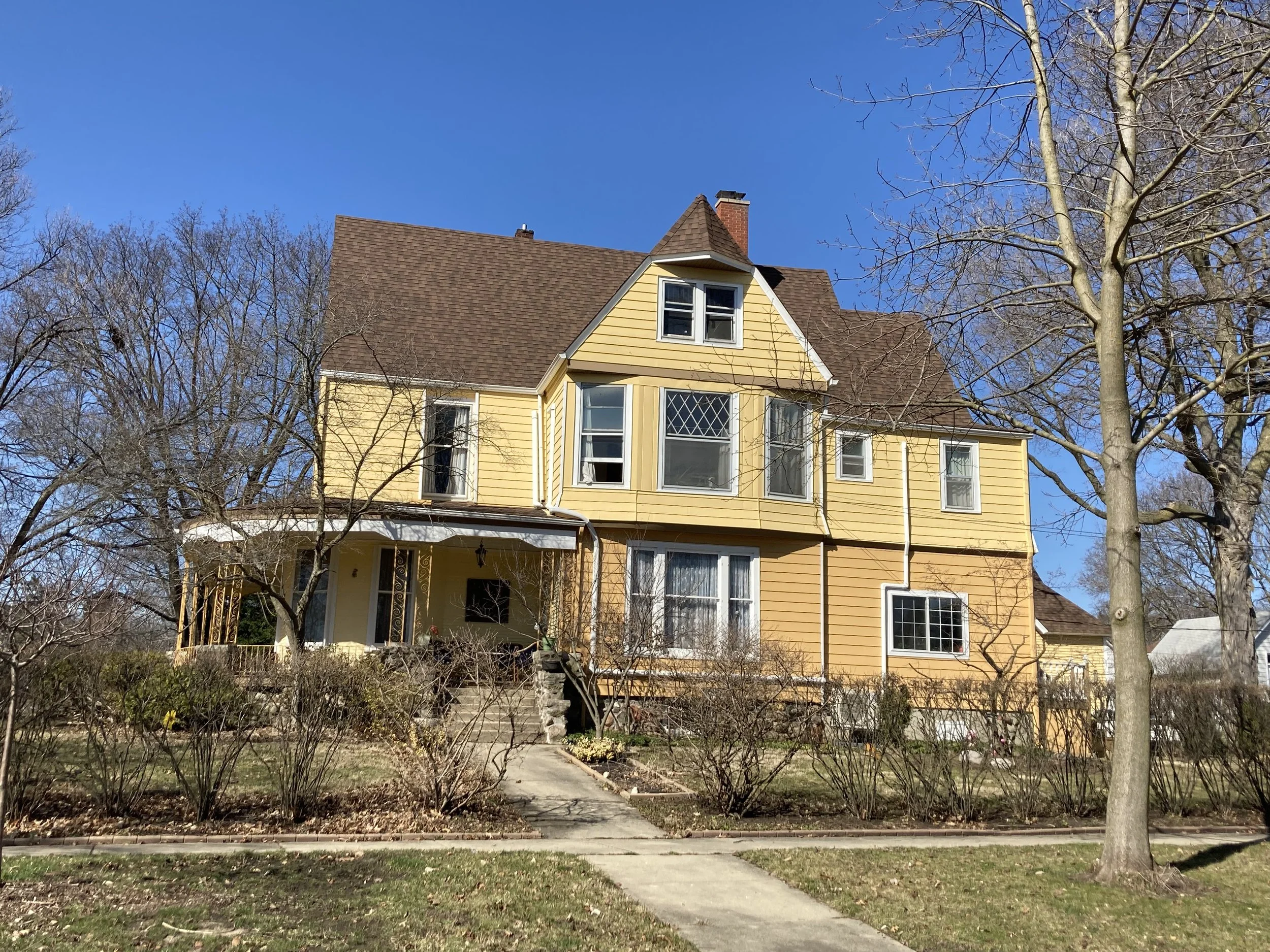 Yellow two-story Victorian-style house with brown roof, front porch, leafless trees, and clear blue sky.