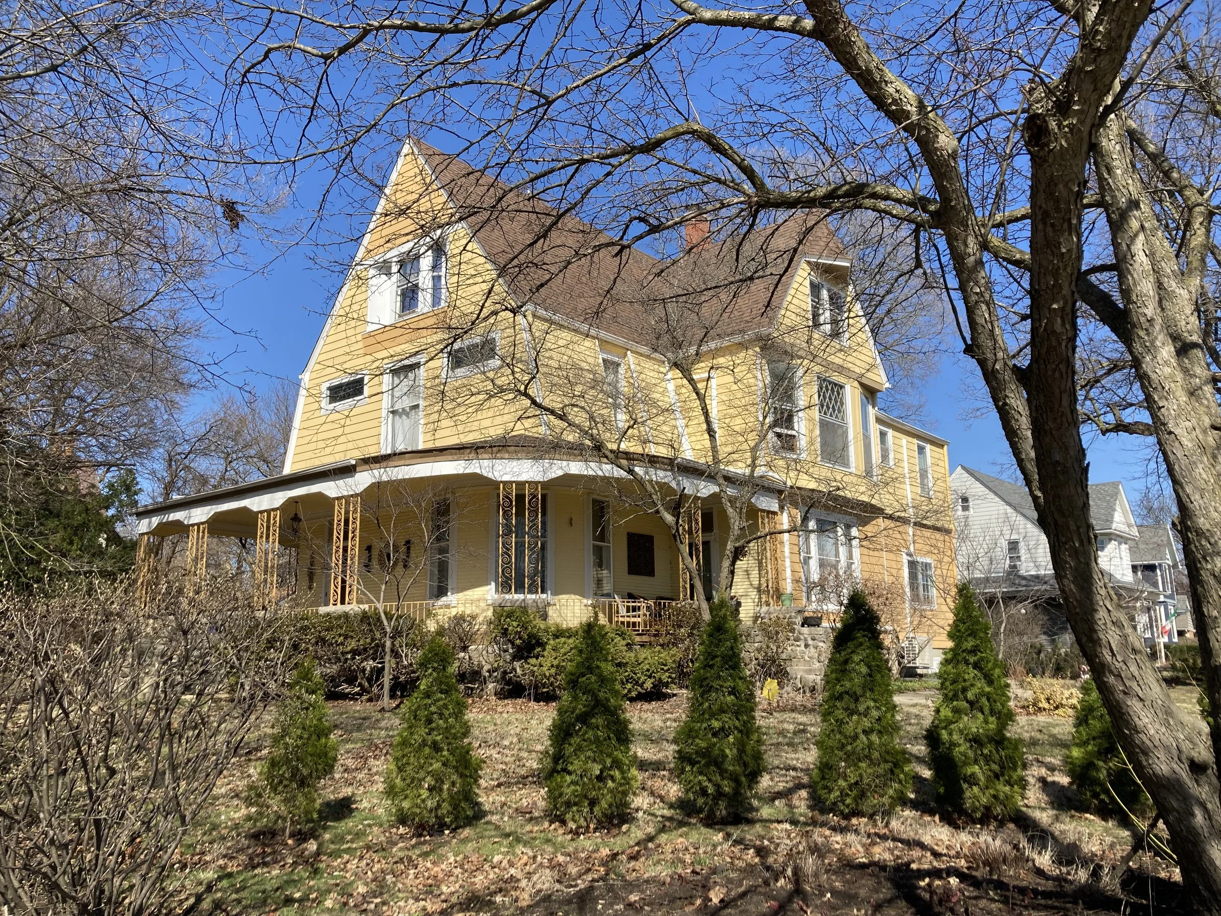 Yellow Victorian-style house with a wraparound porch, leafless trees, and a row of small evergreens under a clear blue sky.