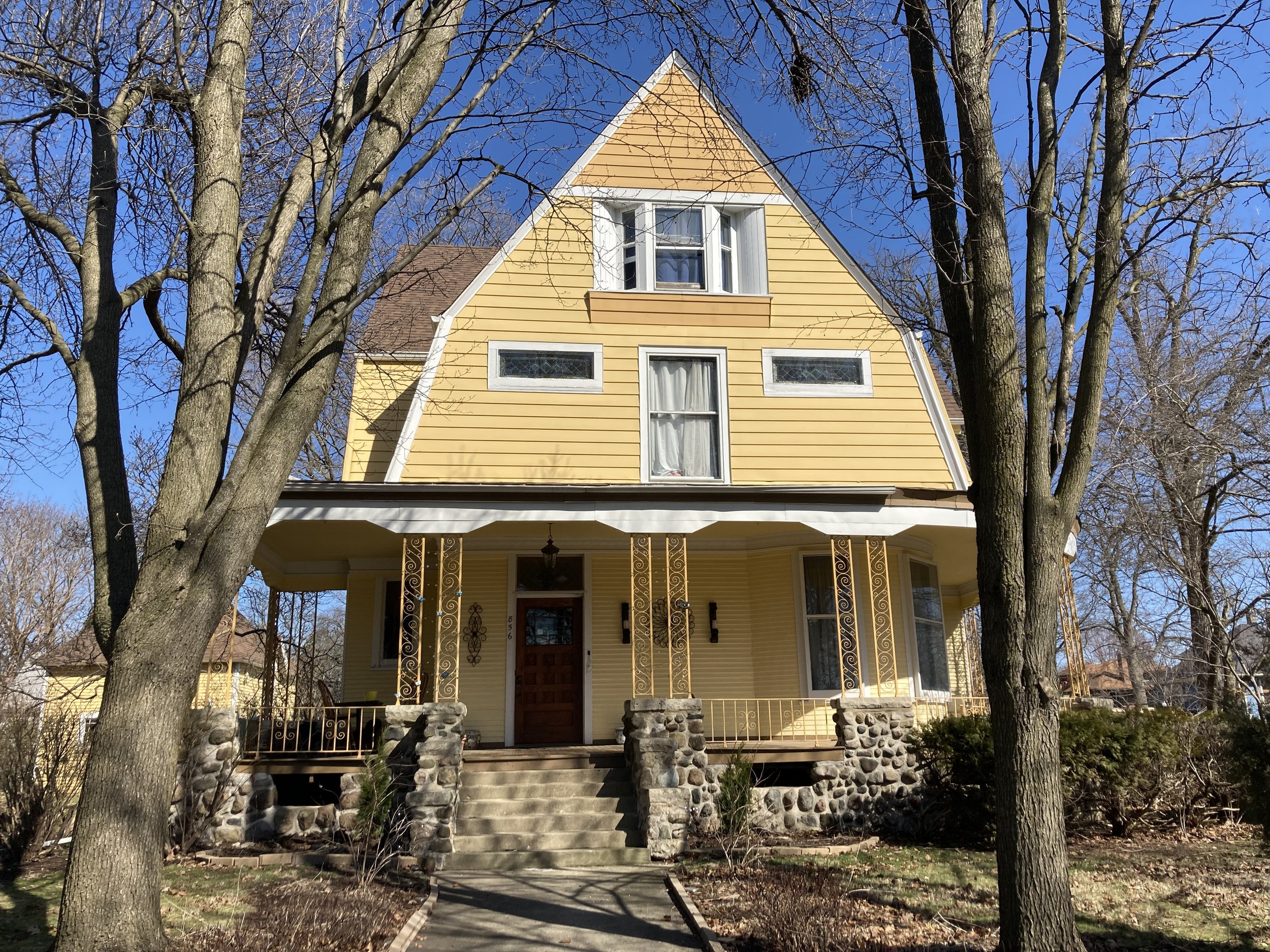 Yellow two-story gable house with a covered front porch, stone foundation, and leafless trees framing it against a clear blue sky.