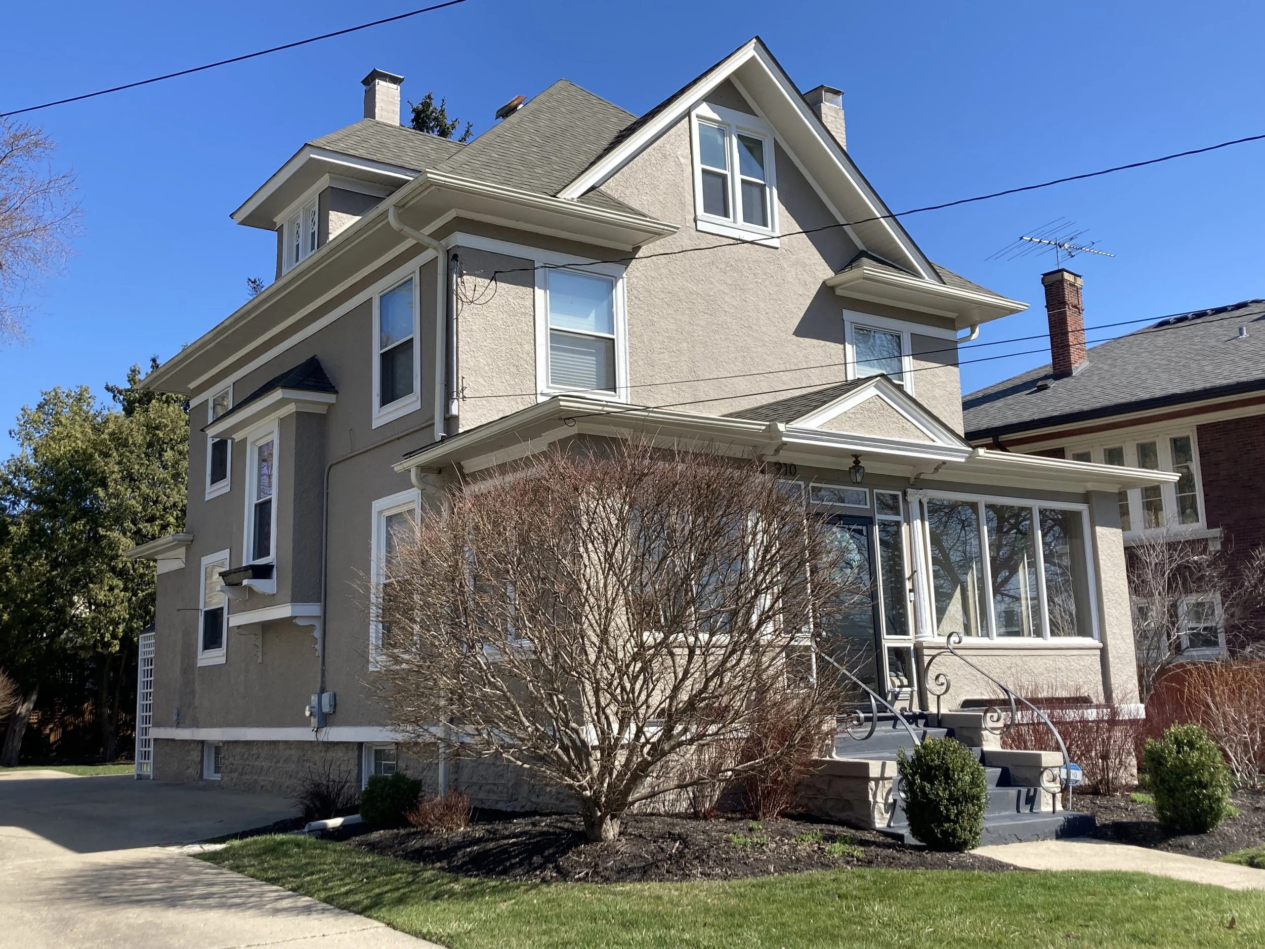 Beige three-story house with enclosed front porch, leafless shrubs, and clear blue sky.