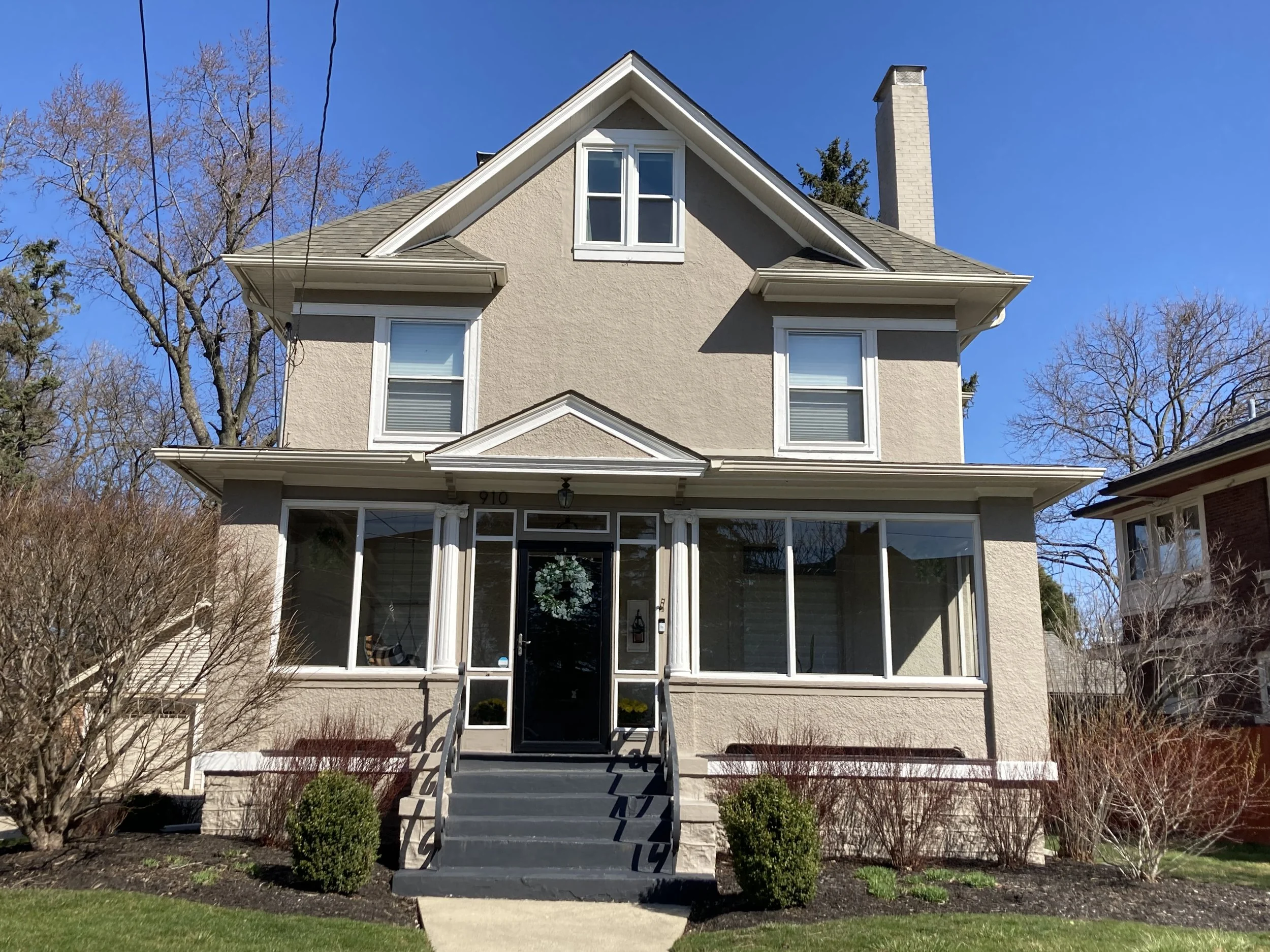 Front view of a two-story beige house with a centered black front door and steps, enclosed front porch windows, trimmed shrubs, and a clear blue sky.