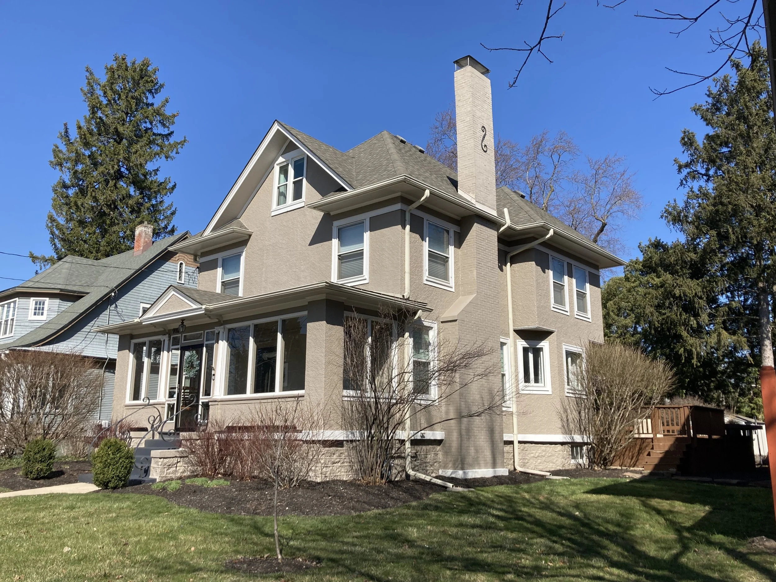 Two-story beige stucco house with a front porch, tall chimney and trimmed lawn under a clear blue sky.