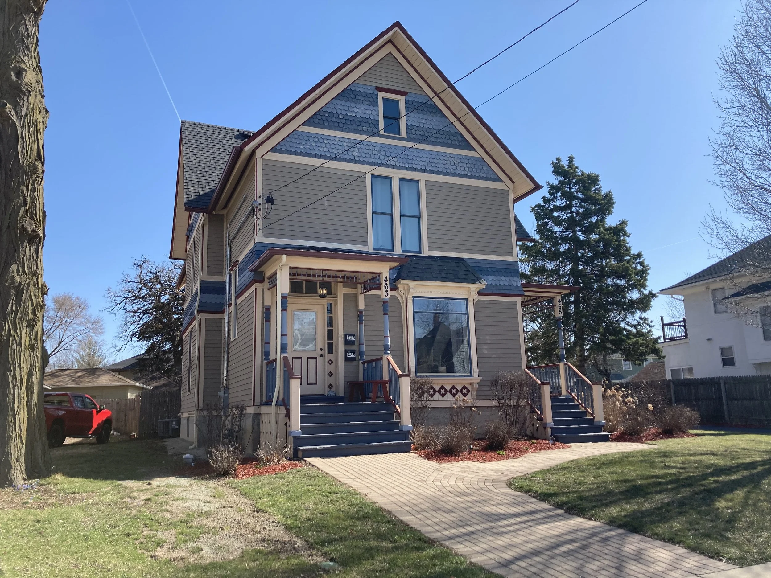 Victorian-style two-story blue-gray house with front porch and walkway, lawn and large trees under a clear blue sky.