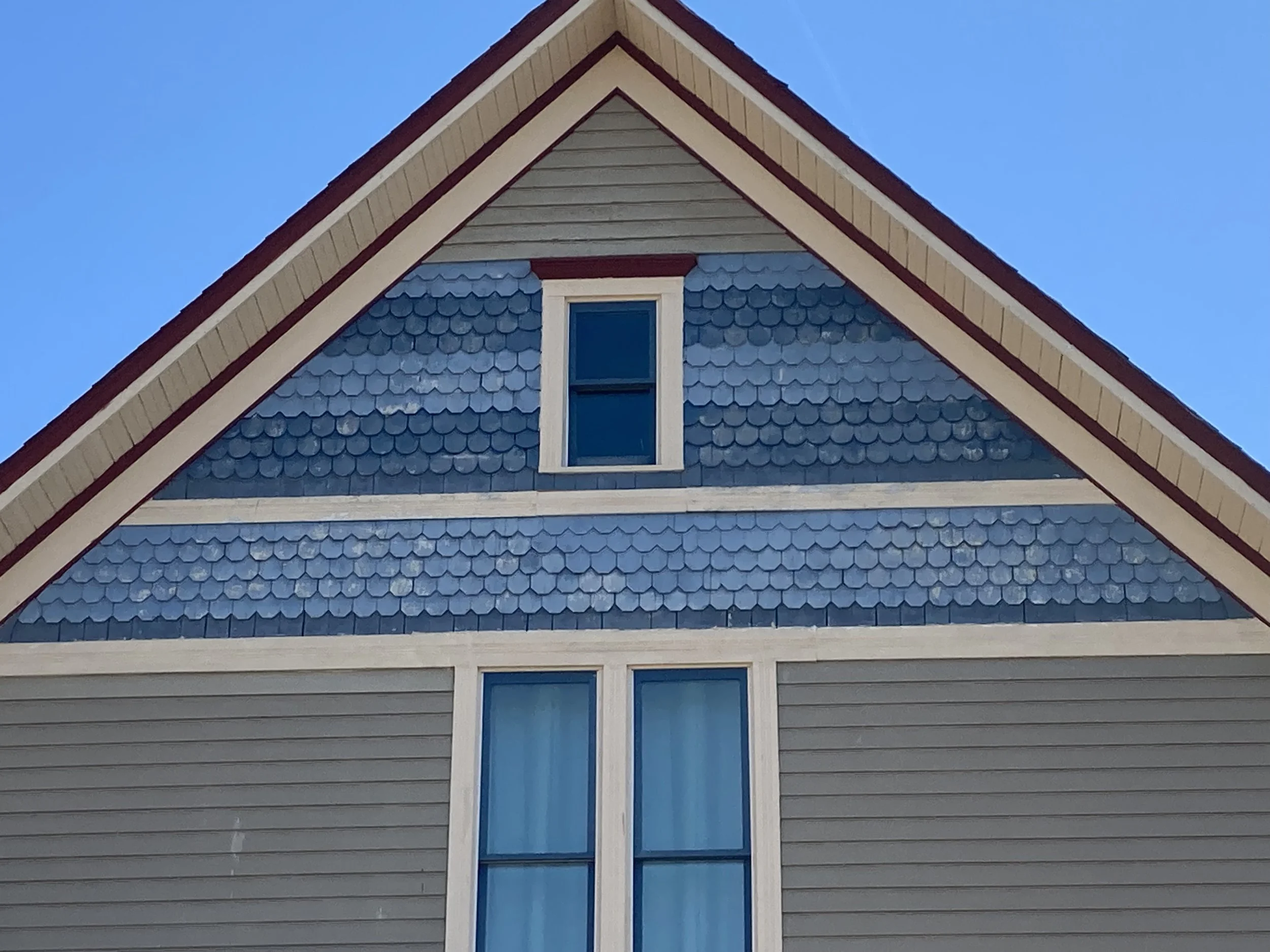 Gable end of a blue-painted house with fish-scale shingles and two white-trimmed windows against a clear blue sky.