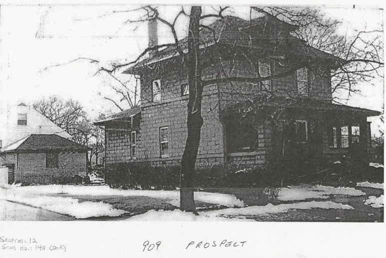 Black-and-white photo of a two-story wood-sided house with a front porch and a leafless tree in the yard, snow patches on the ground, with handwritten "909 PROSPECT" at the bottom.