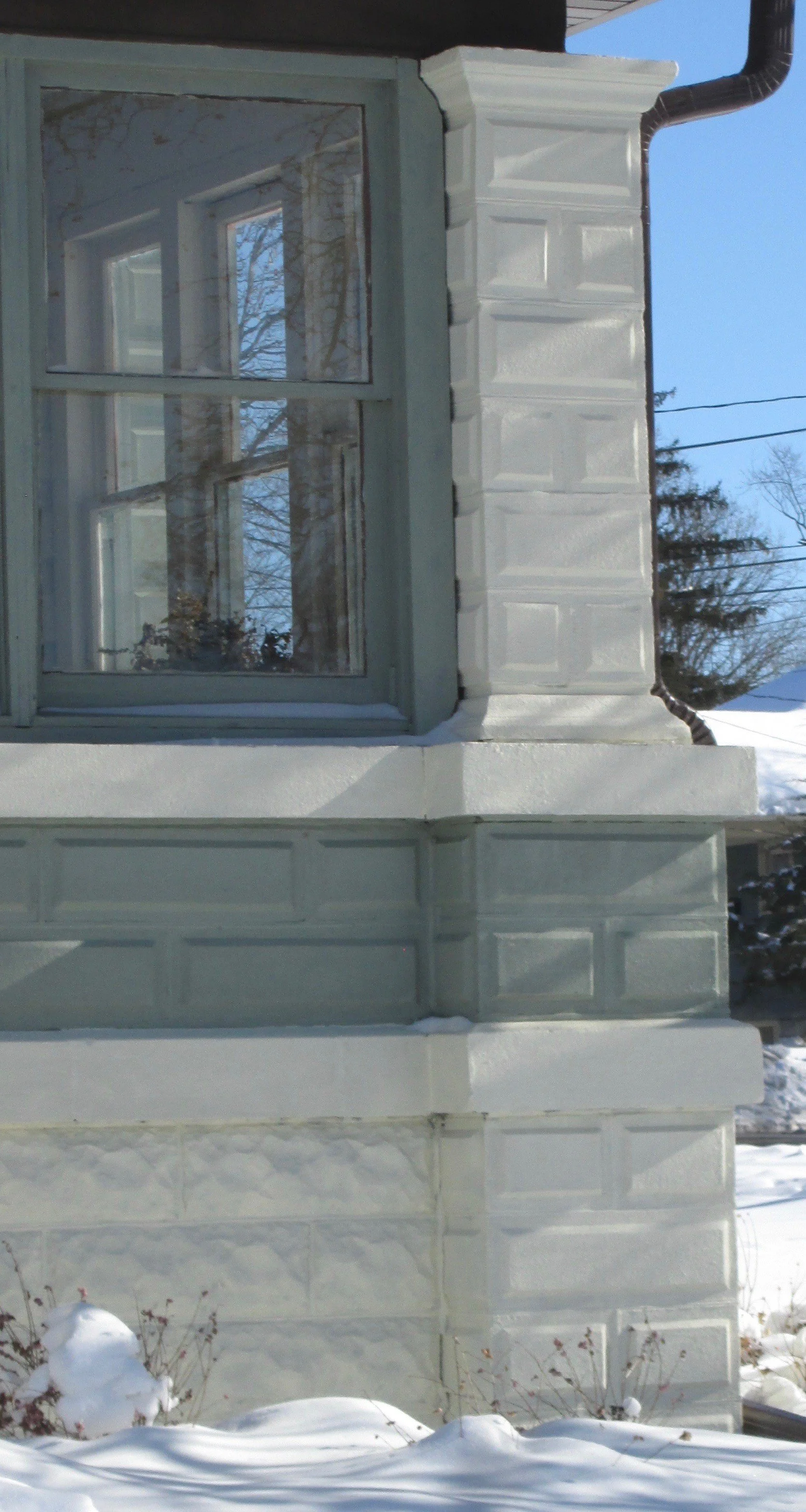 Windowed corner of a pale green and white house with decorative trim and snow at the base.