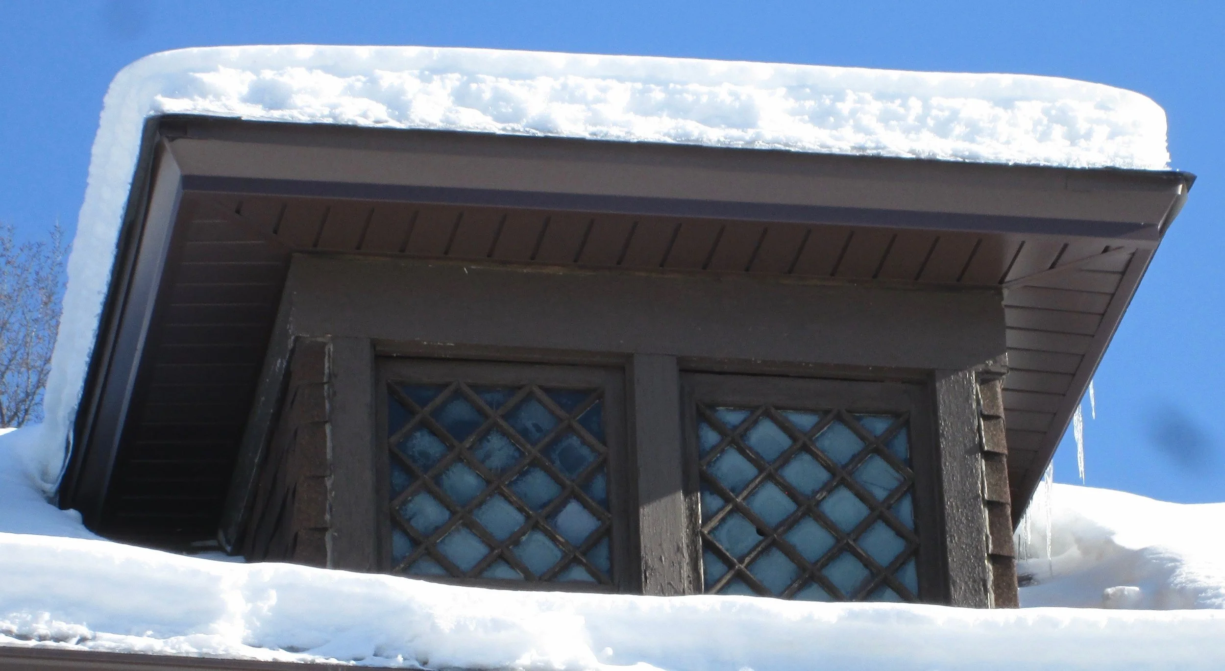 Snow-covered dormer with diamond‑pane windows, icicles hanging from the eaves against a clear blue sky.