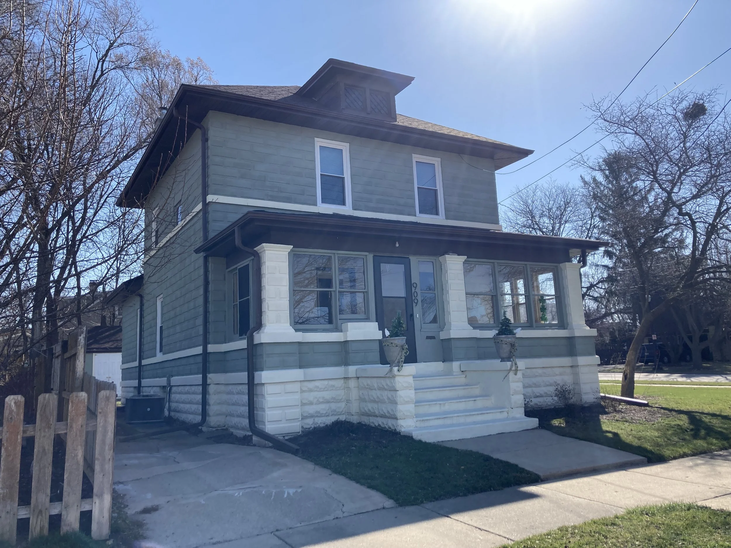 Two-story green house with a covered front porch, white steps and planters, leafless trees and bright sun in a clear blue sky.