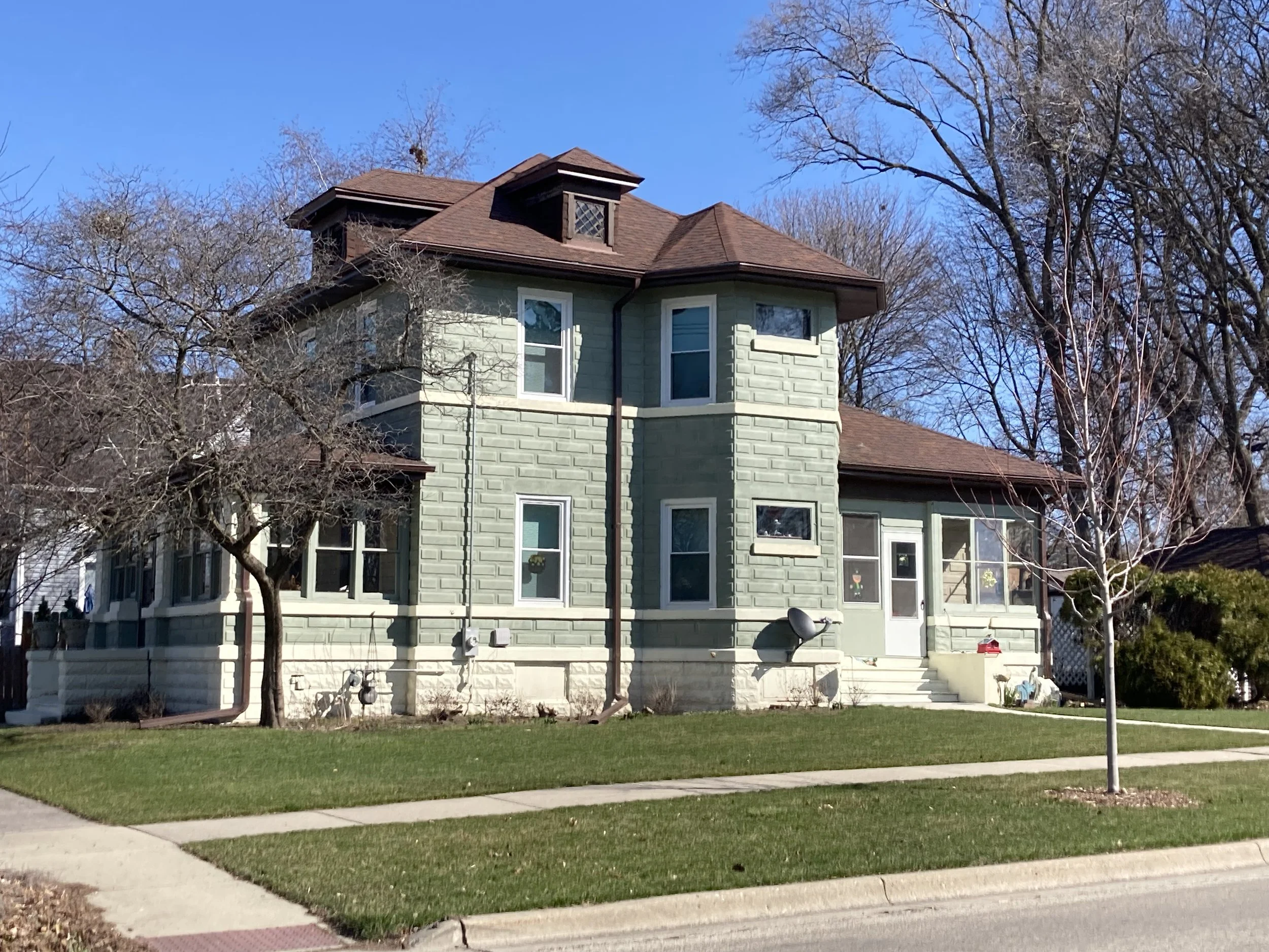 Two-story light green house with a brown roof on a corner lot, leafless trees and a tidy front lawn under a clear blue sky.