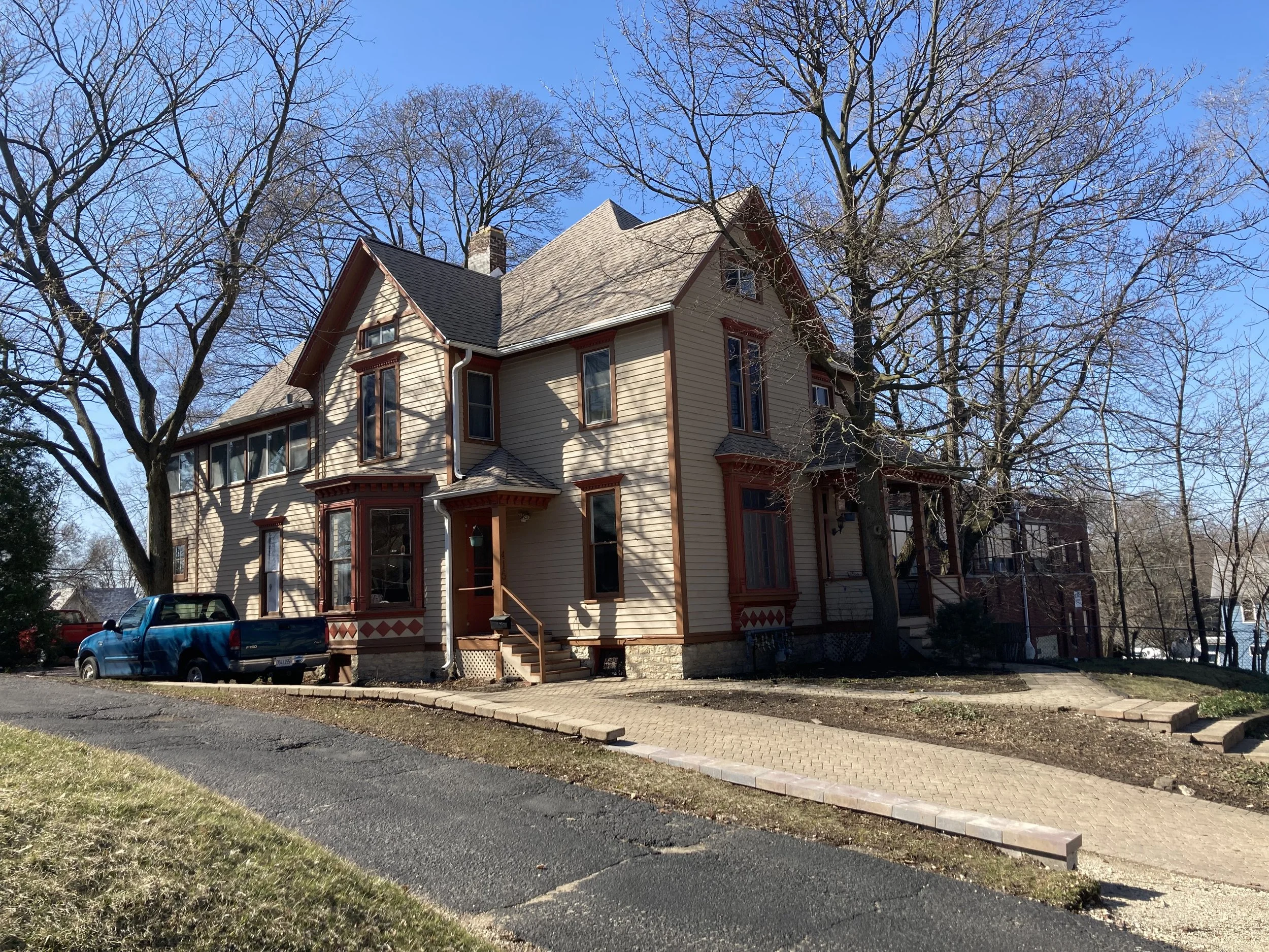 Large two-story Victorian-style beige house with red trim, leafless trees, a blue pickup parked at the side, and a clear blue sky.