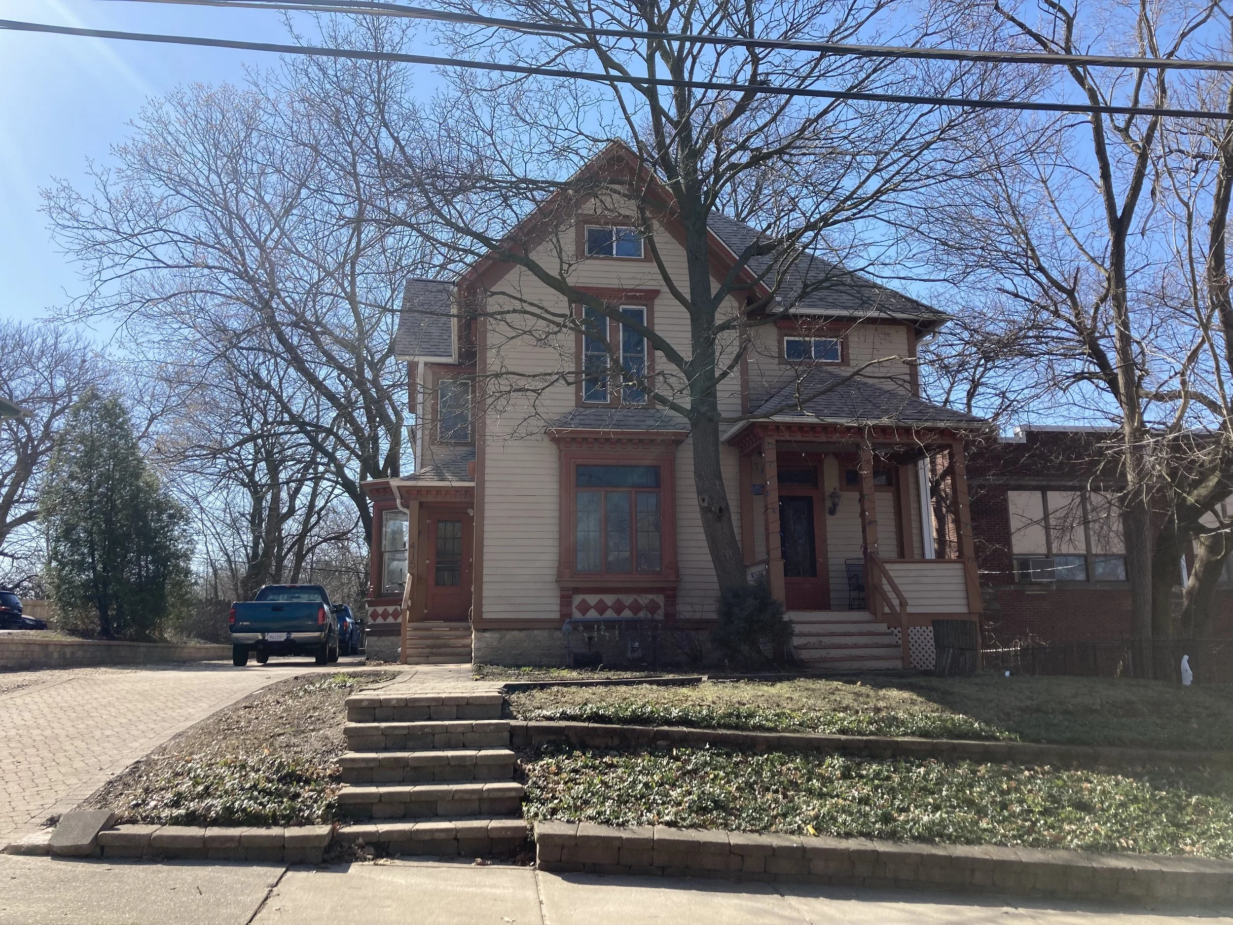 Victorian-style two-story house with a front porch and stone steps, leafless trees around it, a pickup truck in the driveway, and a clear blue sky.