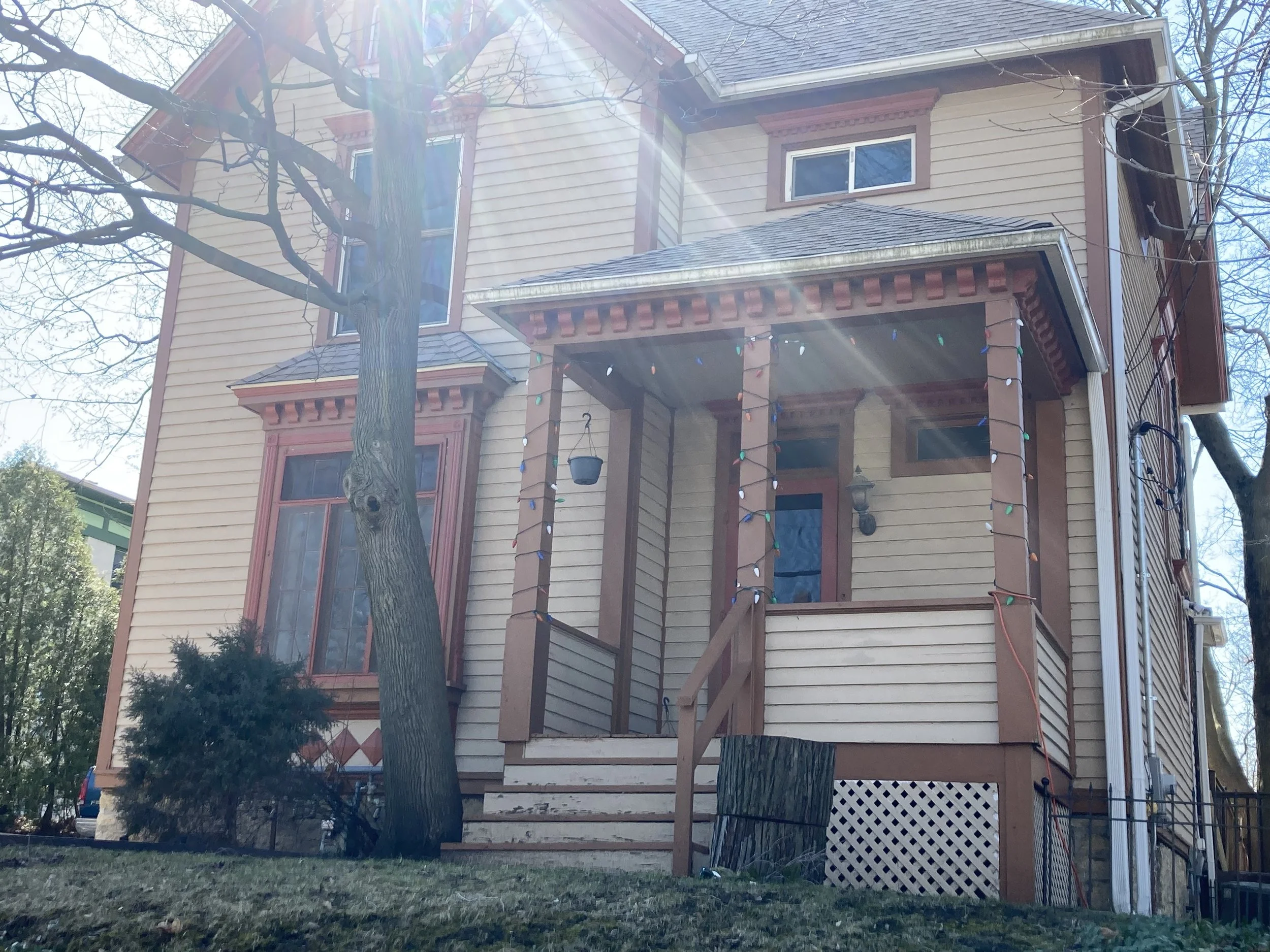 Two-story Victorian-style house with a small covered front porch, bare trees and a leaning trunk in front, multicolored string lights, and sun glare from above.