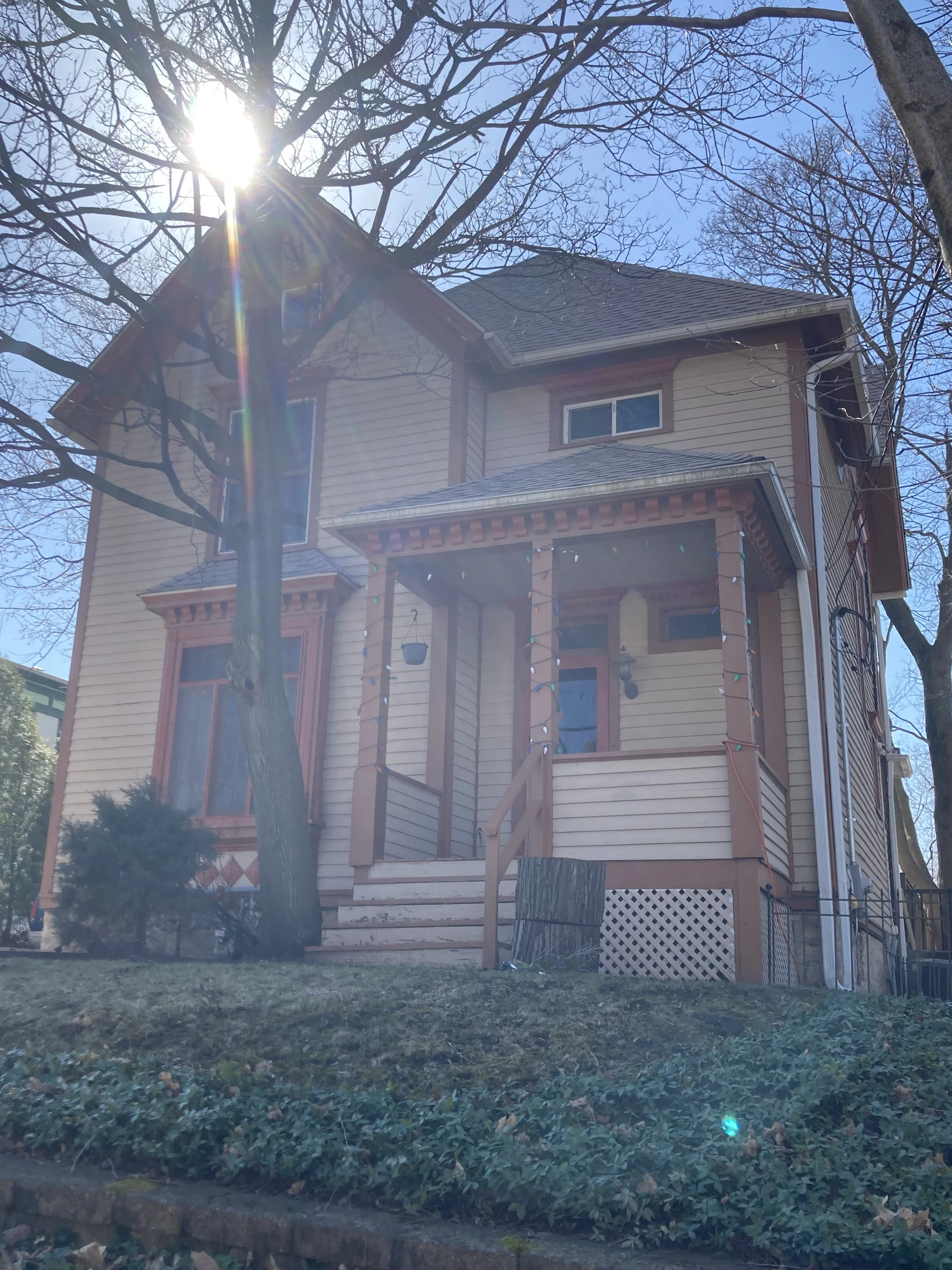 Two-story pale-yellow Victorian house with a front porch, bare trees and sun glare.